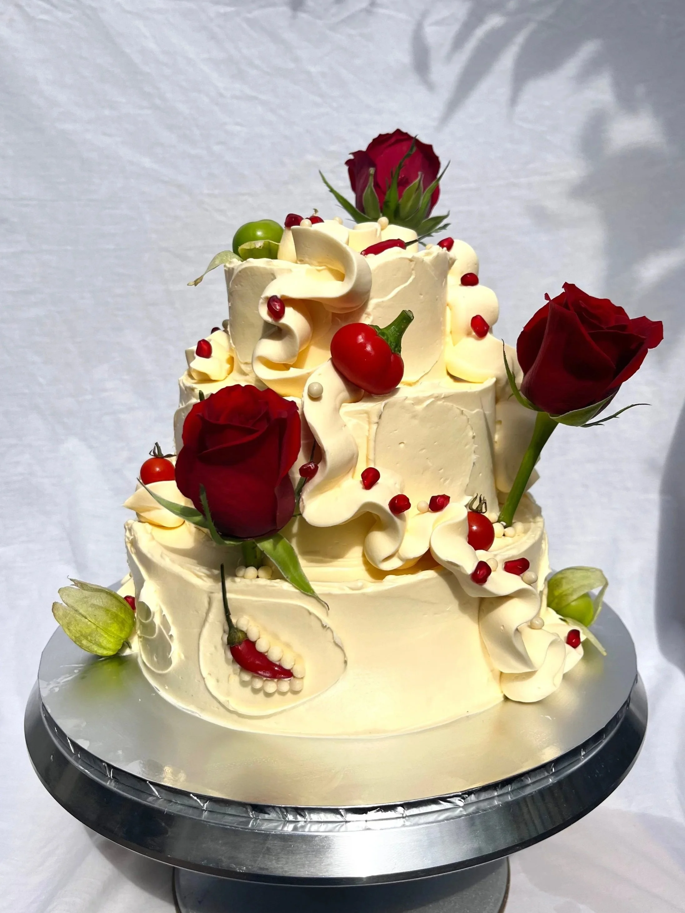 A multi-tiered cake decorated with red roses, green tomatoes, red chillies, white chocolate crispy pearl, displayed on a silver cake stand.