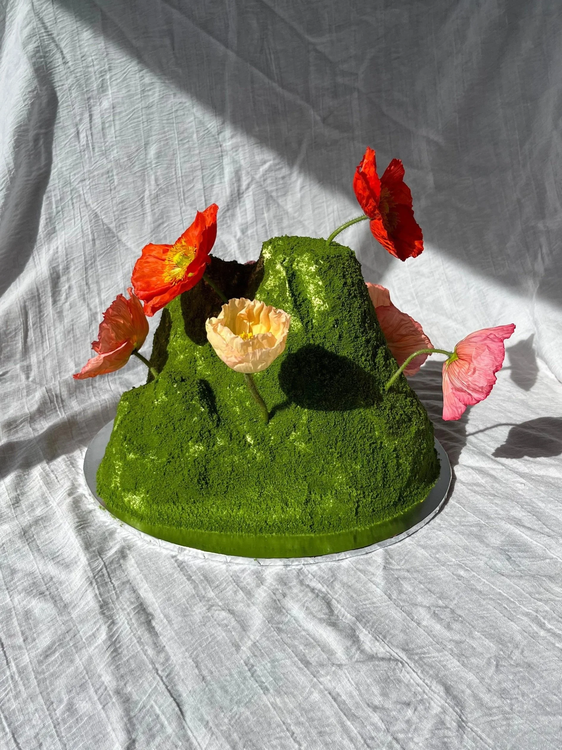 A sculptural cake resembling a mound of bright green moss with colourful poppies, displayed on a round mirror on a light-colored fabric surface.