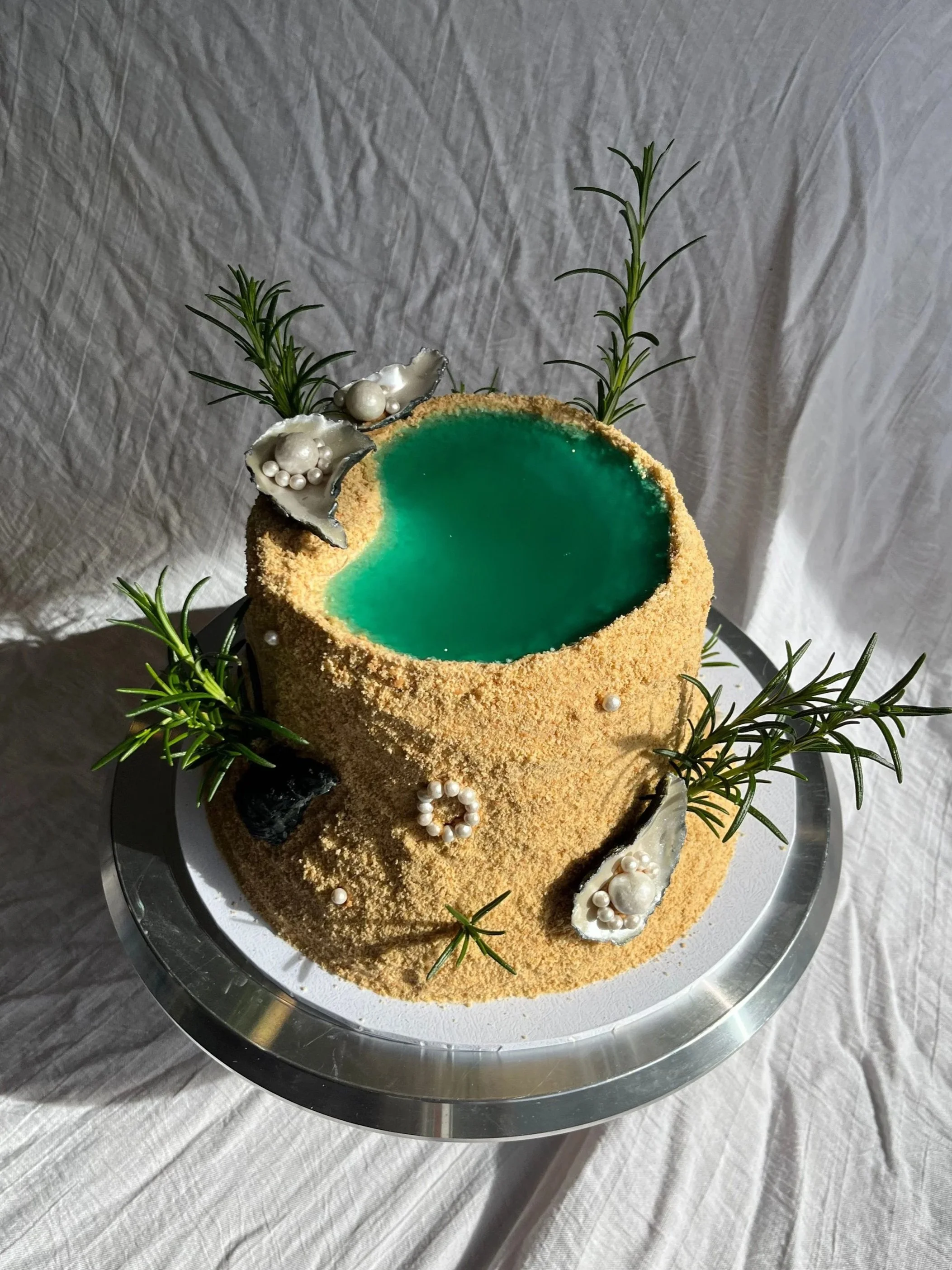A cake made to look like an oasis with edible biscuit sand, an aqua jelly rockpool, edible chocolate oyster shells and  decorated with white edible pearls and silver accents, on a silver tray.