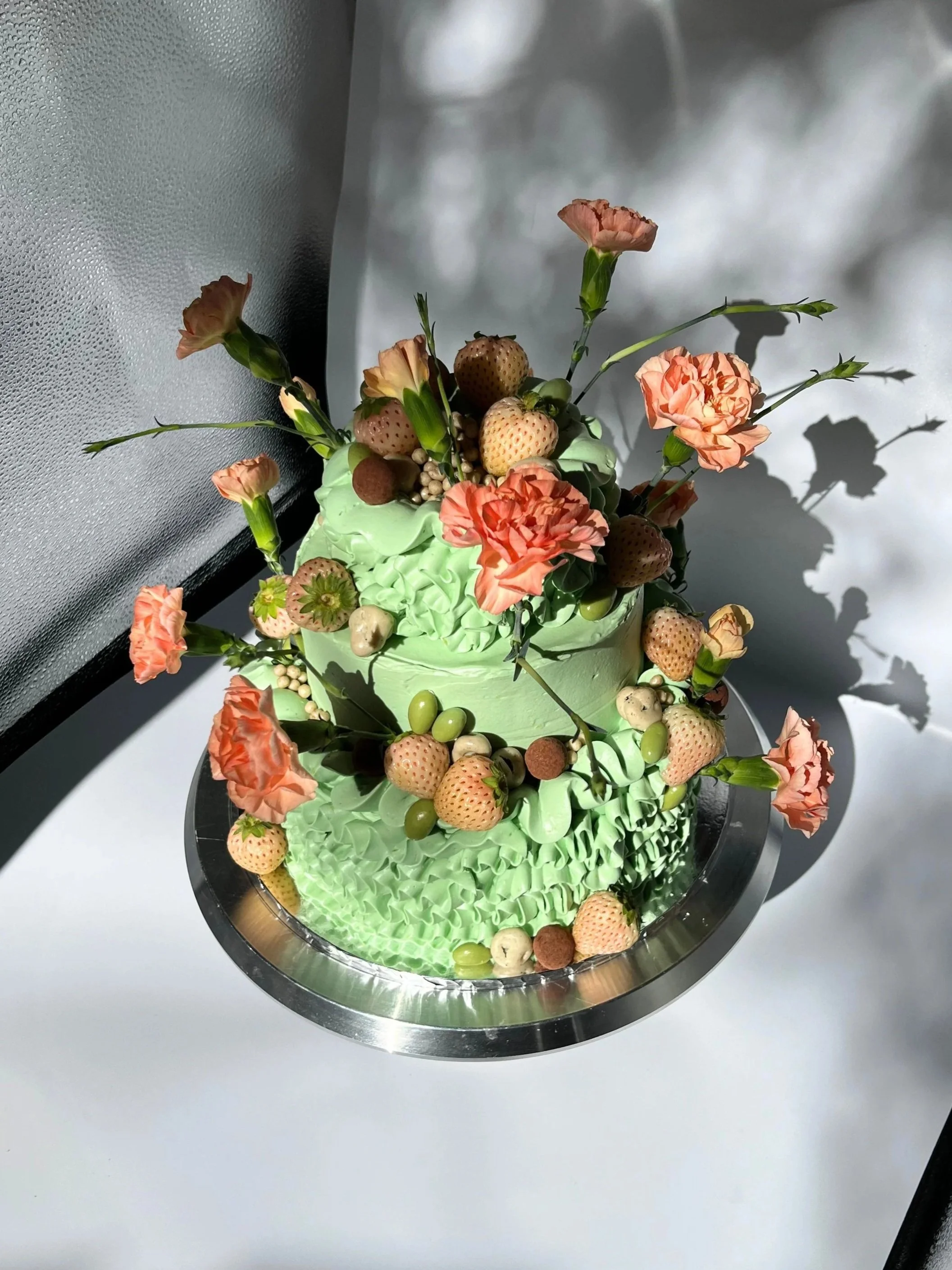 A vibrant mint green cake decorated with pink carnations and an assortment of white strawberries and chocolate rocks, placed on a round reflective metallic cake stand.
