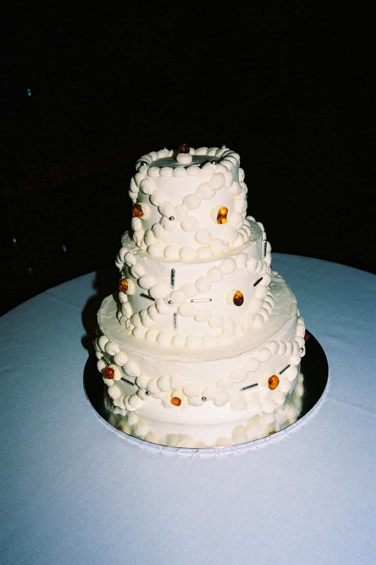 Four-tiered white wedding cake decorated with buttercream piping, amber gemstones, edible silver beads, placed on a round table with a white tablecloth.