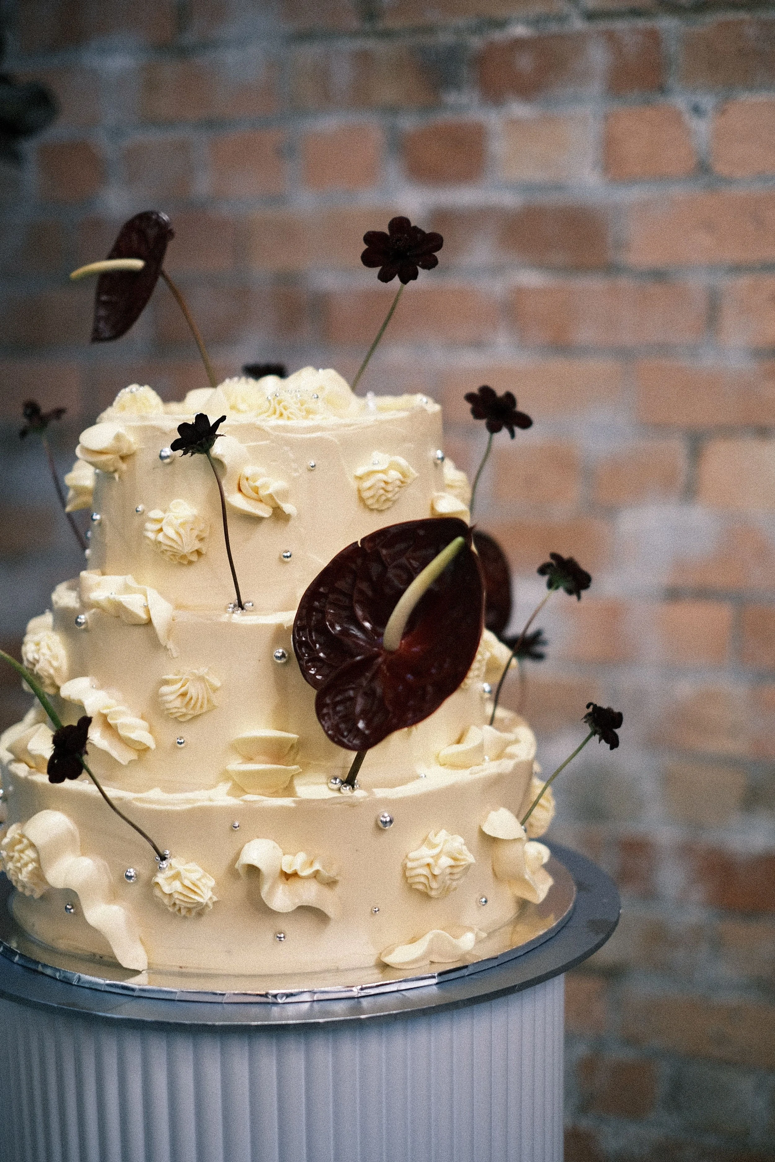 A multi-tiered wedding cake decorated with white frosting, dark chocolate cosmos, dark red anthuriums, and small silver and white decorative balls, set against a brick wall background.