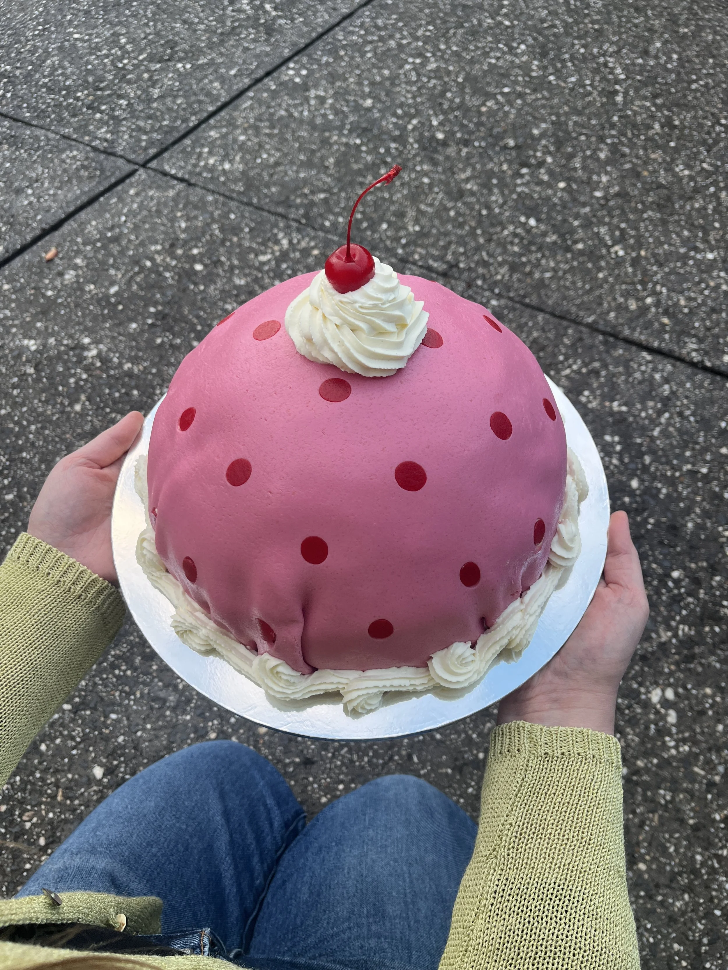 Person holding a large, pink polka dot Princess cake. Covered in pink and red polka dot marzipan and decorated with a cherry and whipped cream on top, sitting on a sidewalk.