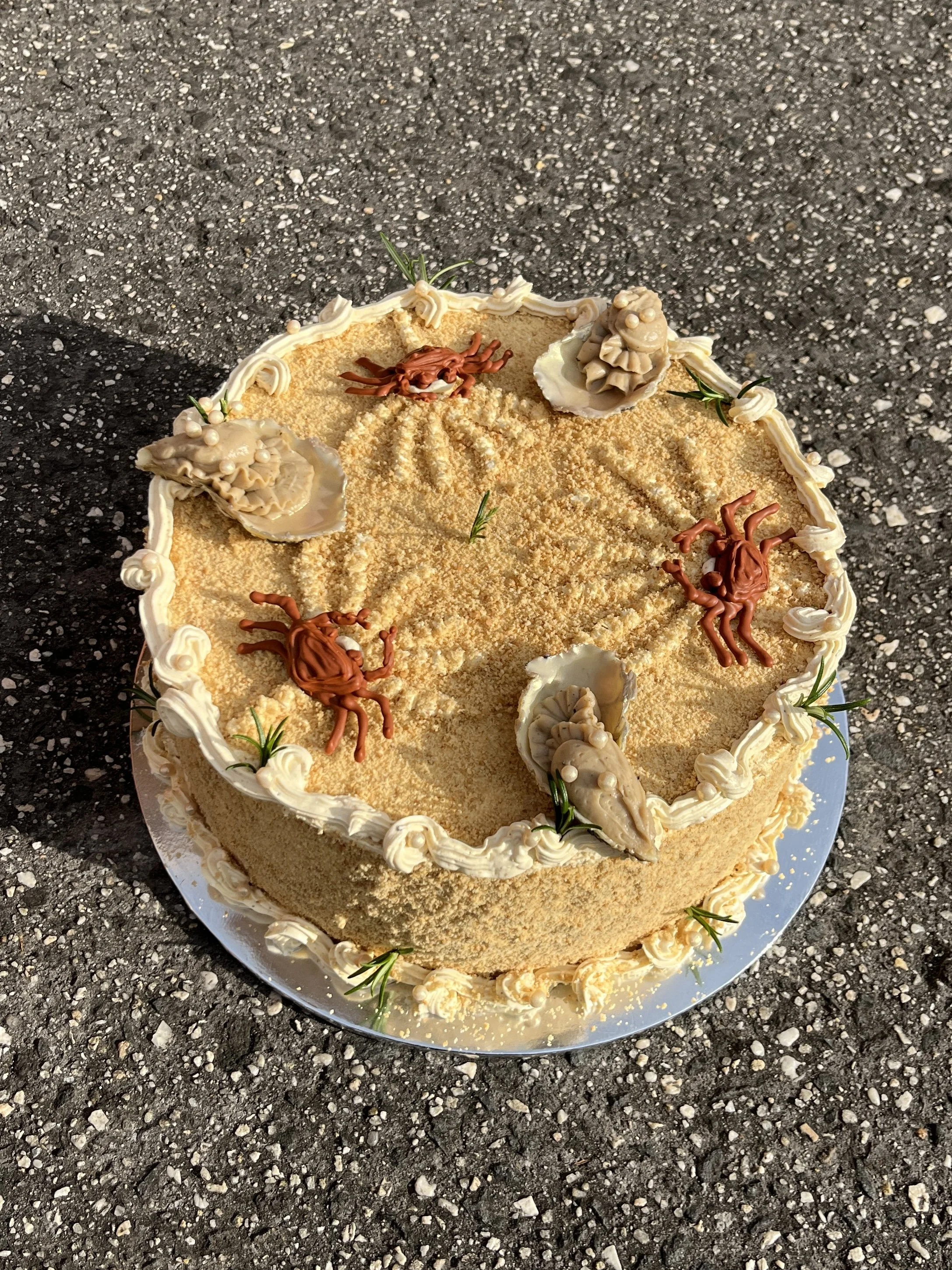 A round cake decorated with small crab and shell toppers, rosemary, and whipped cream on a silver base, placed on a rough asphalt surface.