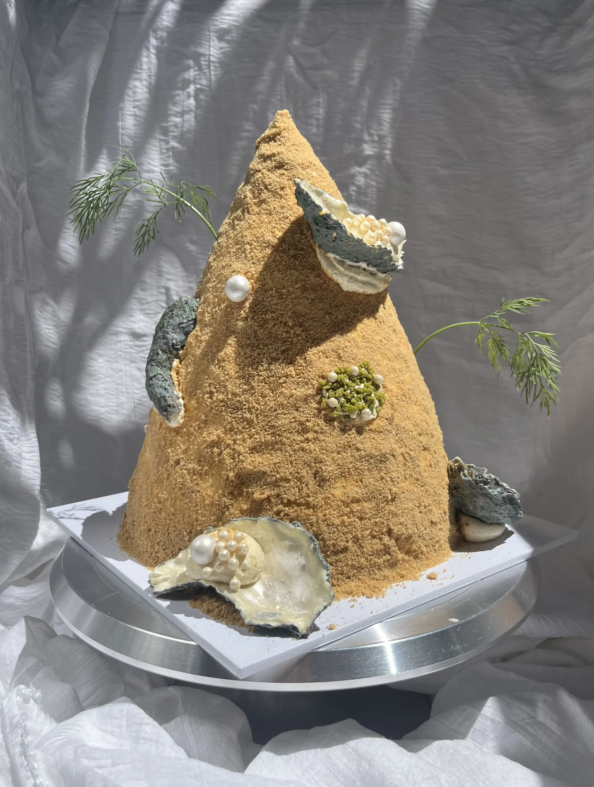 A cake made to look like a sandcastle, decorated with white and pearl-colored edible beads, edible chocolate oyster shells, and green leafy accents, placed on a silver platter.