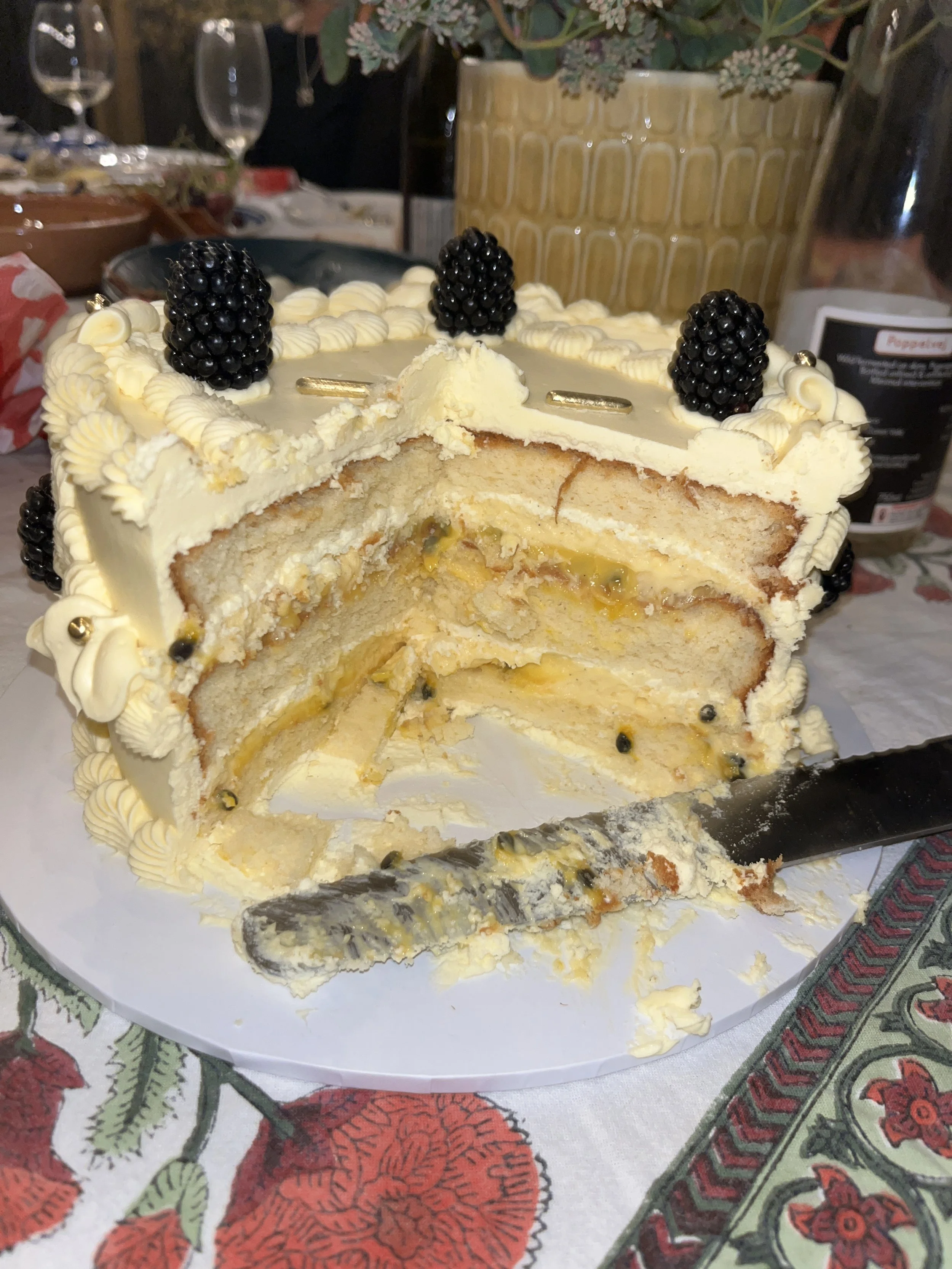 A partially cut and eaten vanilla chiffon cake with passionfruit curd, cream cheese frosting and blackberries on top, decorated with piping, golden sprinkles, sitting on a white cake board on a festive table with a floral-patterned tablecloth.