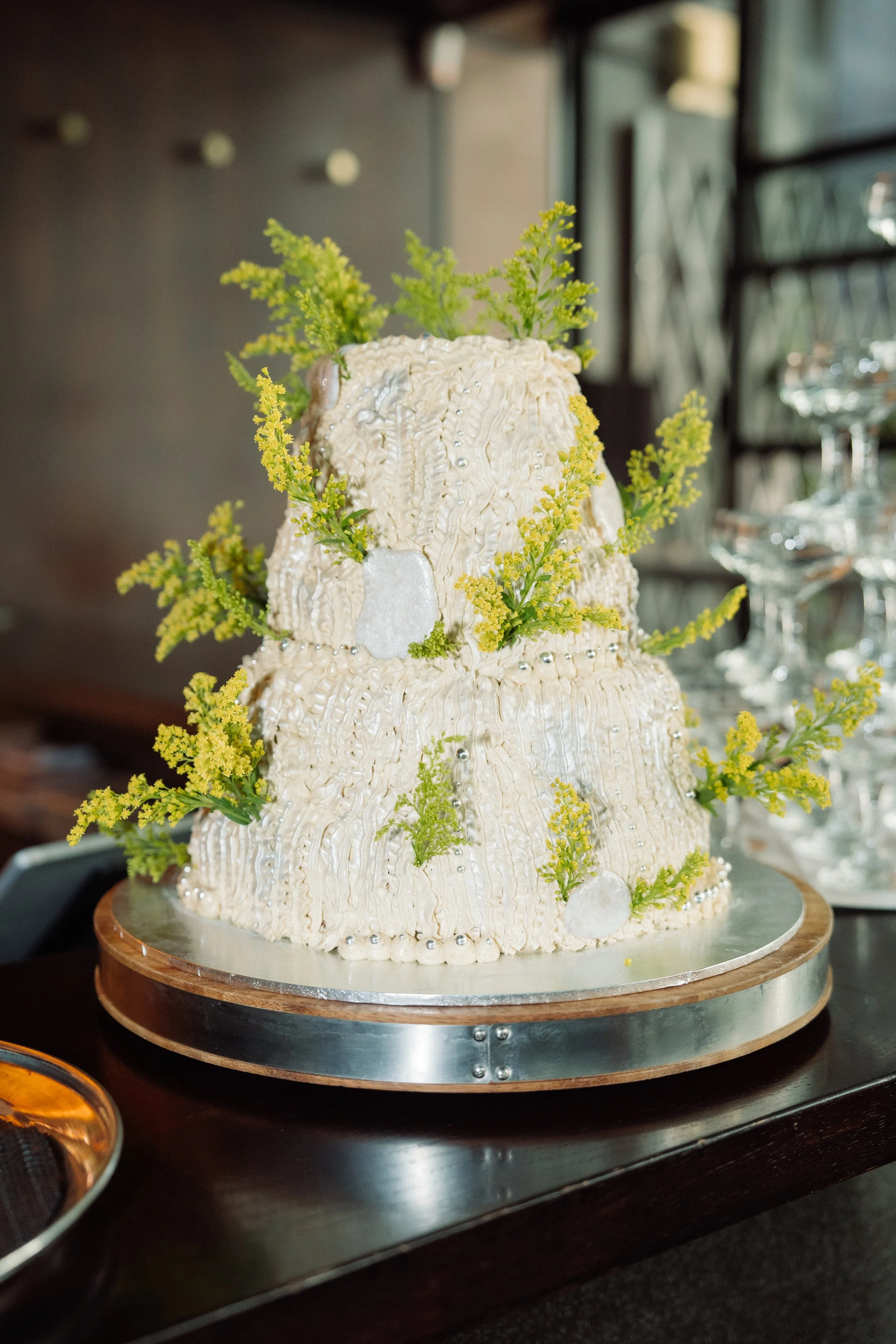 A tiered wedding cake decorated with cream-colored icing, small edible silver balls, and golden rod flowers, displayed on a wooden and silver stand.