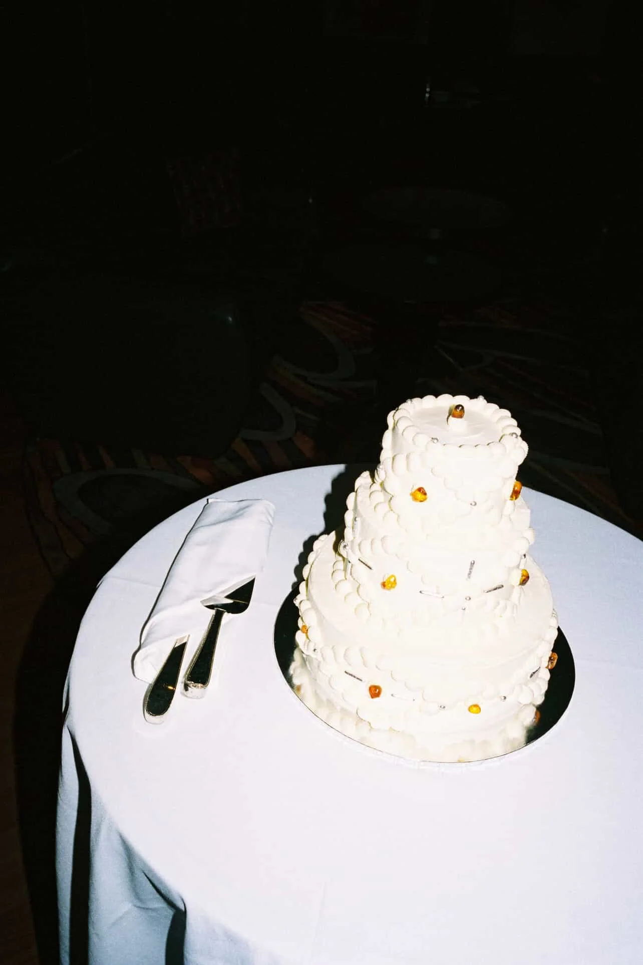 A four-tiered white wedding cake decorated with small amber gemstones, placed on a round white table with a white cloth. There are cake serving utensils on the table beside the cake.