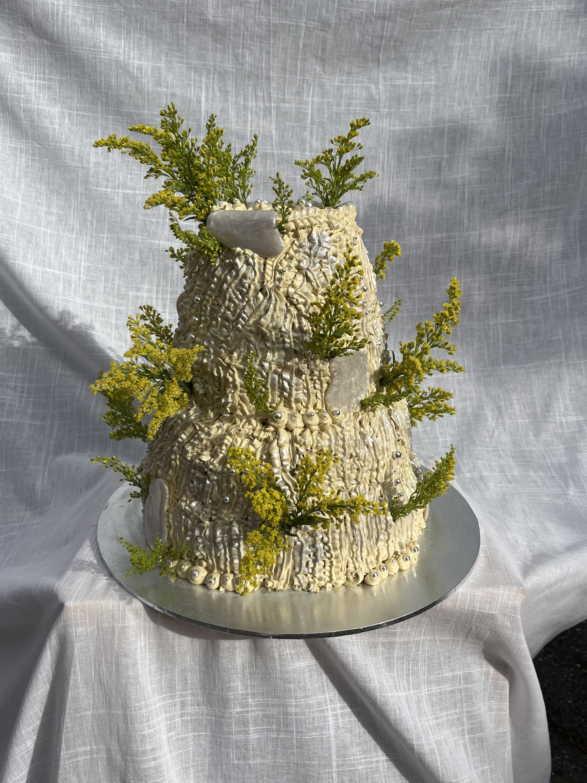 Multi-tiered cake decorated with cream cheese buttercream, edible silver pearls, and golden rod flowers, set on a round silver cake board