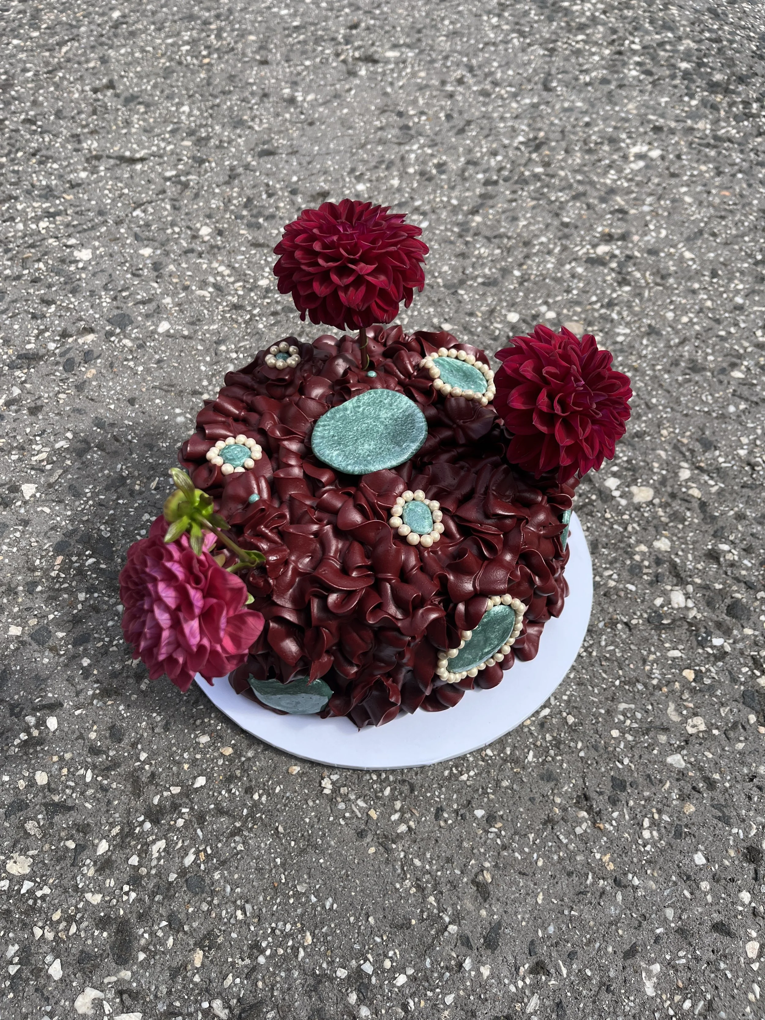 A round cake resembling a flower bouquet, made of deep maroon petal-like piping with some turquoise embellished edible stones and edible pearl beads, placed on a white circular base on a rough asphalt surface.