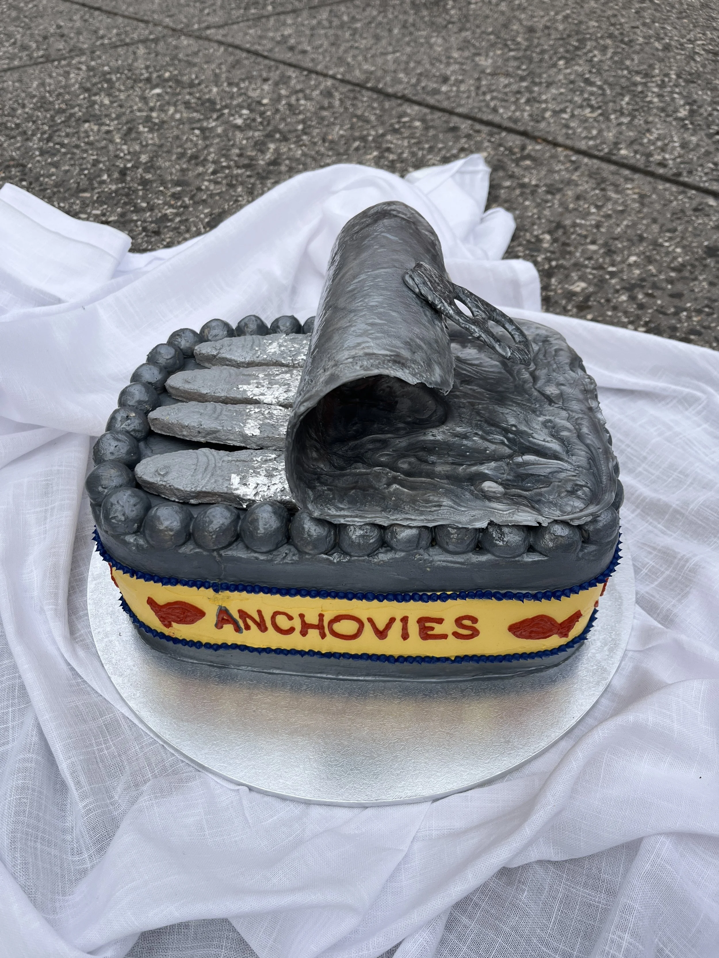 A cake shaped like an anchovy tin with the front decorated with the words 'ANCHOVIES' and two fish images, surrounded by a beaded border, placed on a white cloth.