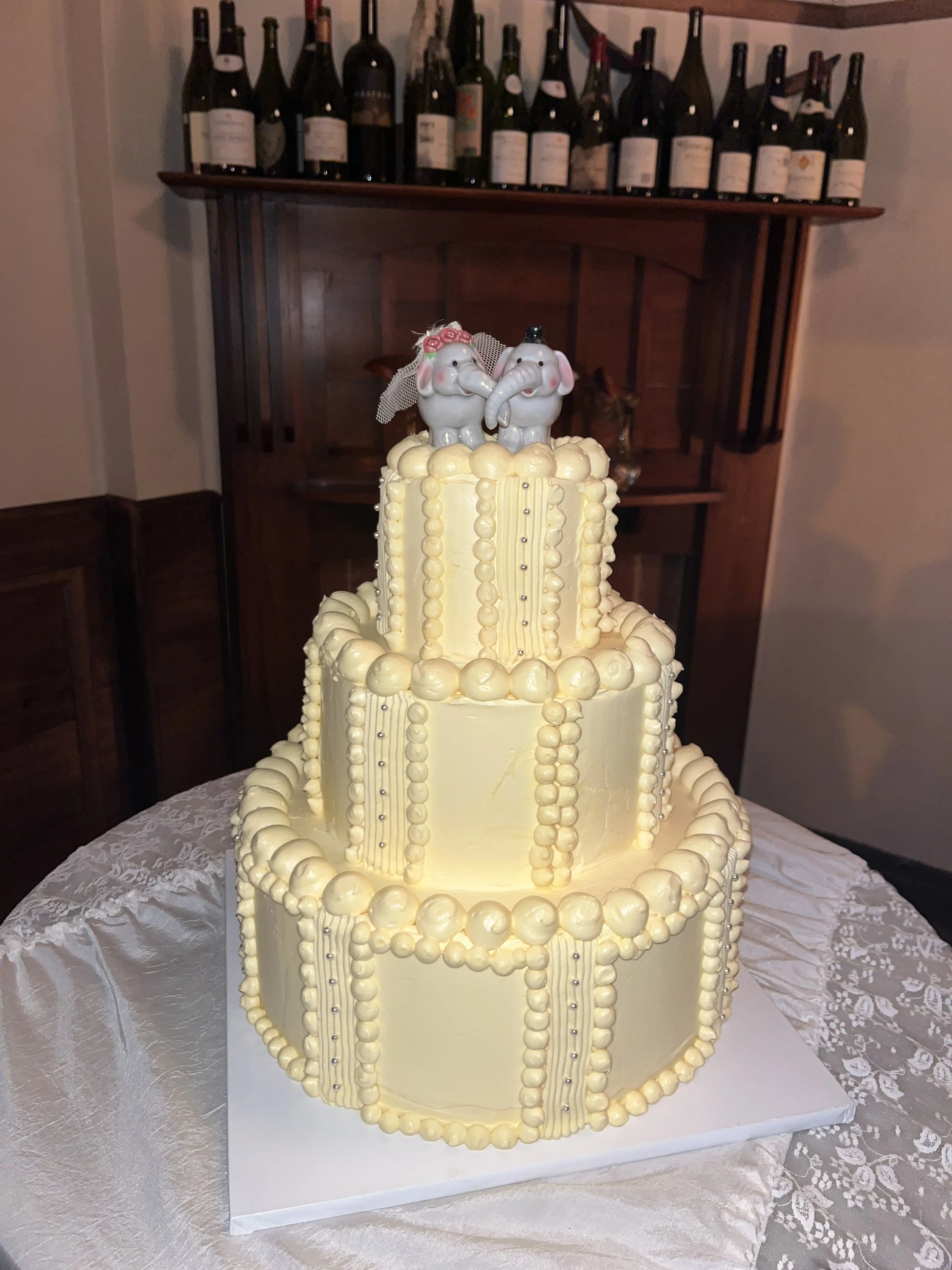 A three-tiered wedding cake. The top of the cake features two elephant figurines dressed as a bride and groom, with the bride elephant wearing a veil and pink roses, and the groom elephant wearing a top hat.