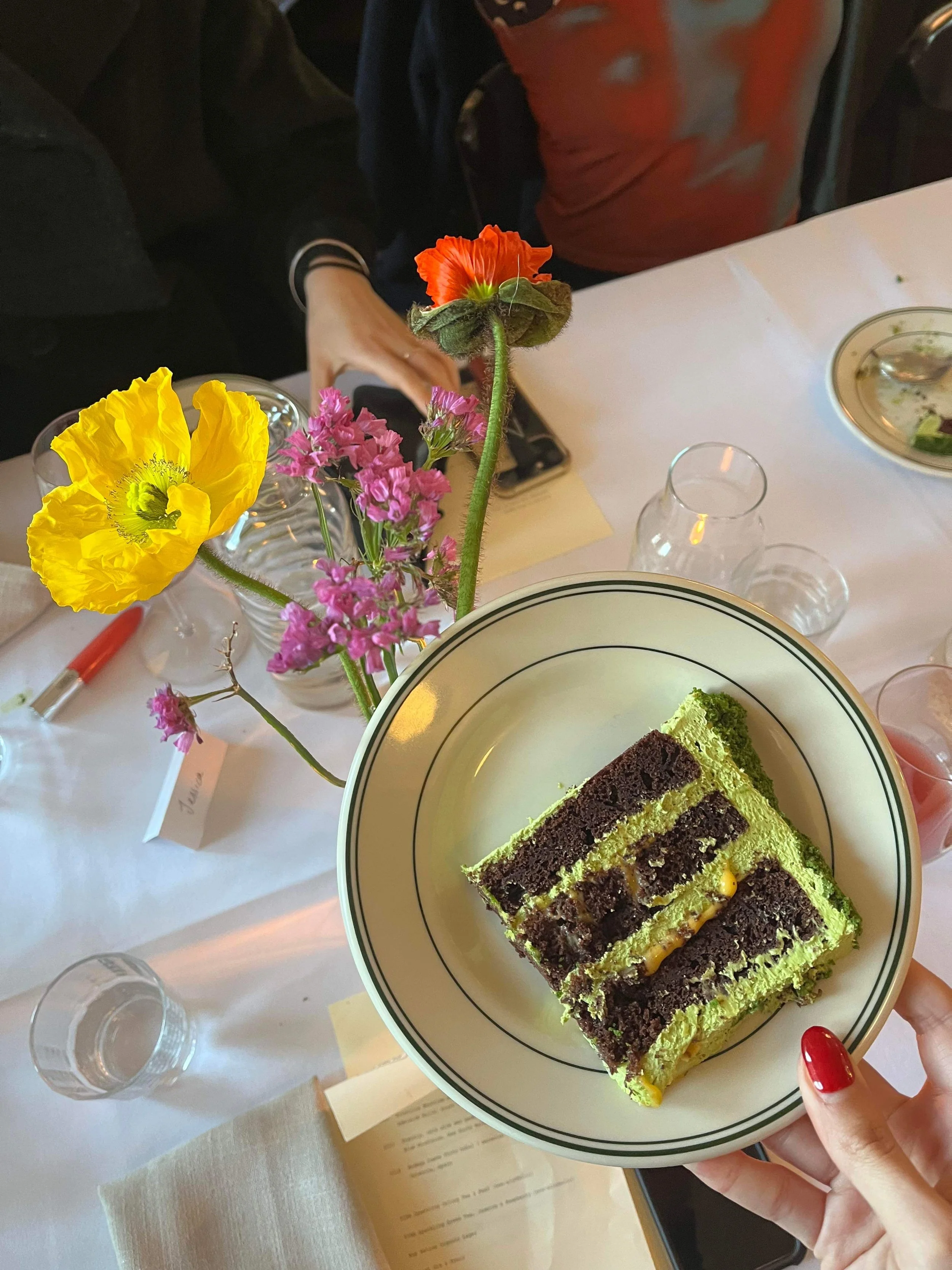 A white plate with slices of layered matcha and chocolate cake with  passionfruit curd, held by a person's hand with red-painted nails. In the background, a table with a vase of poppies, drinking glasses, and a dessert plate.