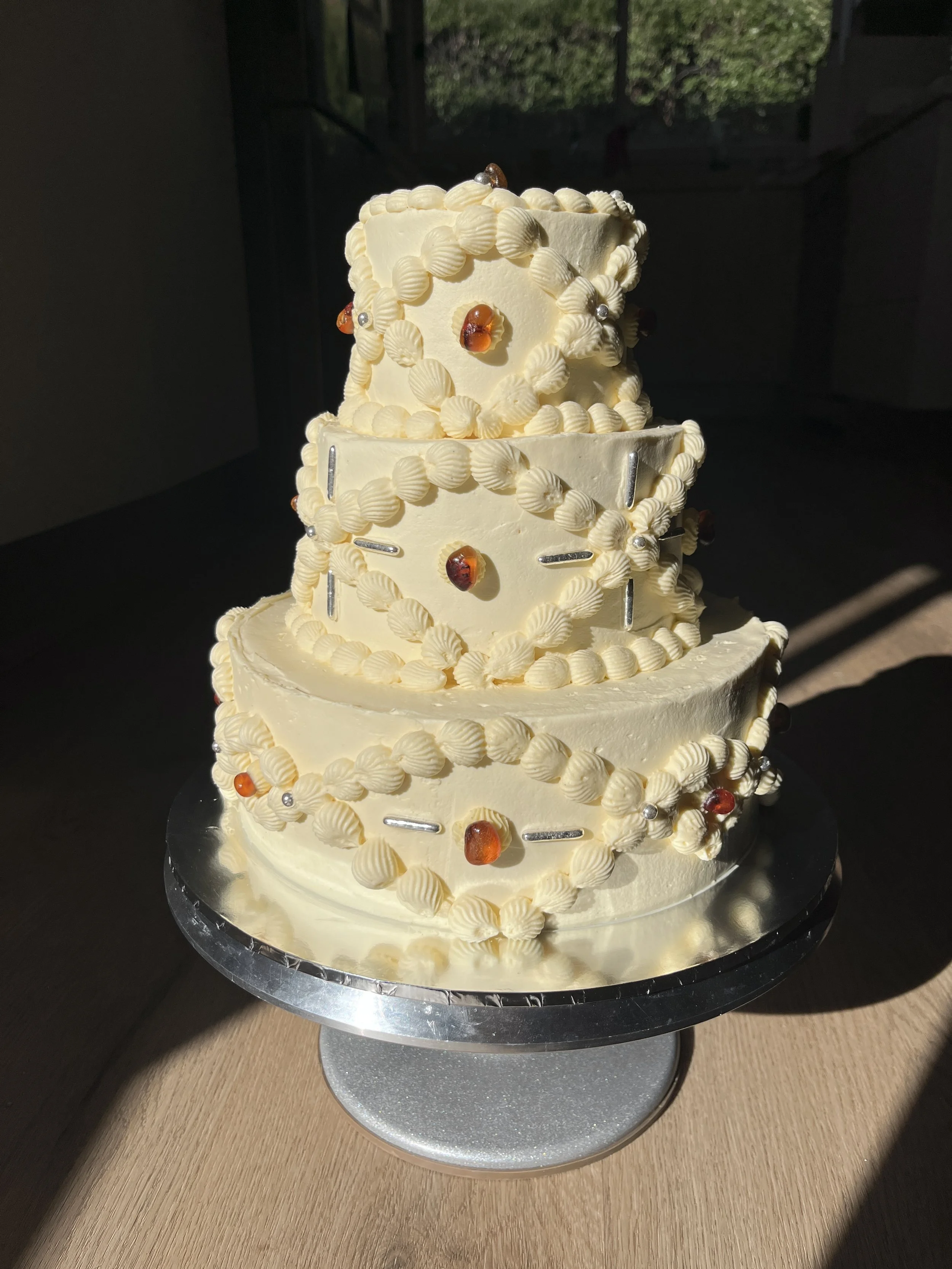 A multi-layered wedding cake decorated with white buttercream, amber beads, and silver dragees, placed on a silver cake stand on a wooden surface with sunlight casting shadows.