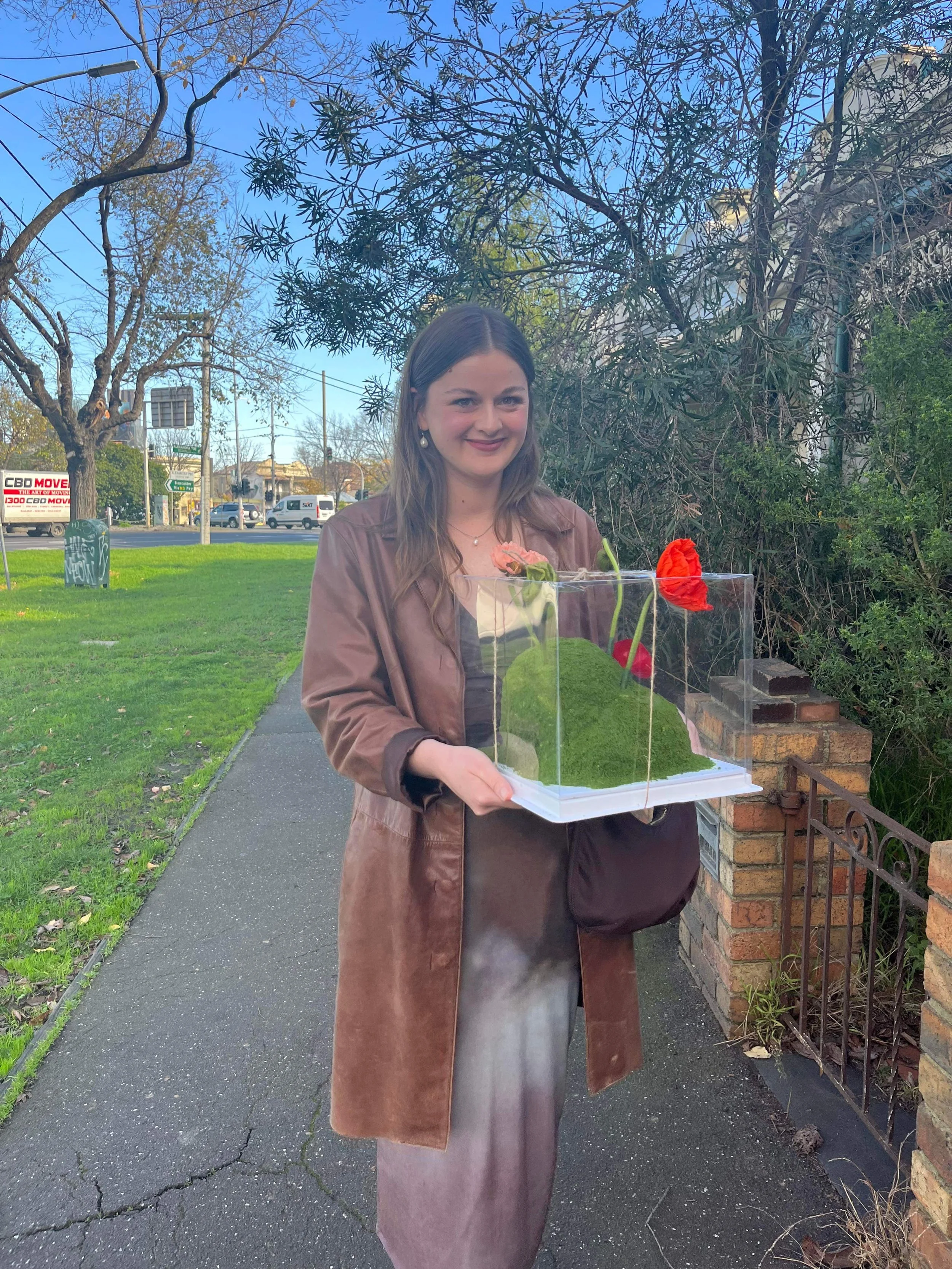 A woman standing on a sidewalk holding a transparent cake box with a floral decorated cake inside. She is wearing earrings, a brown coat, and a grey dress, smiling at the camera.