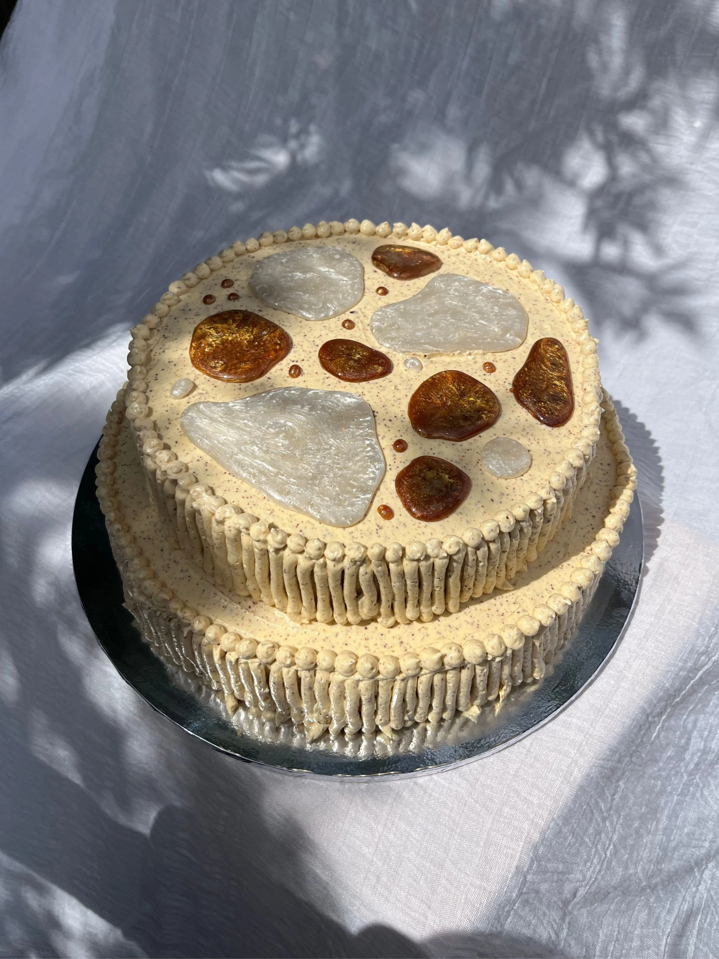 A round cake with brown butter frosting decorated with large clear and amber-colored edible stones on top, placed on a silver cake board and metallic tablecloth.