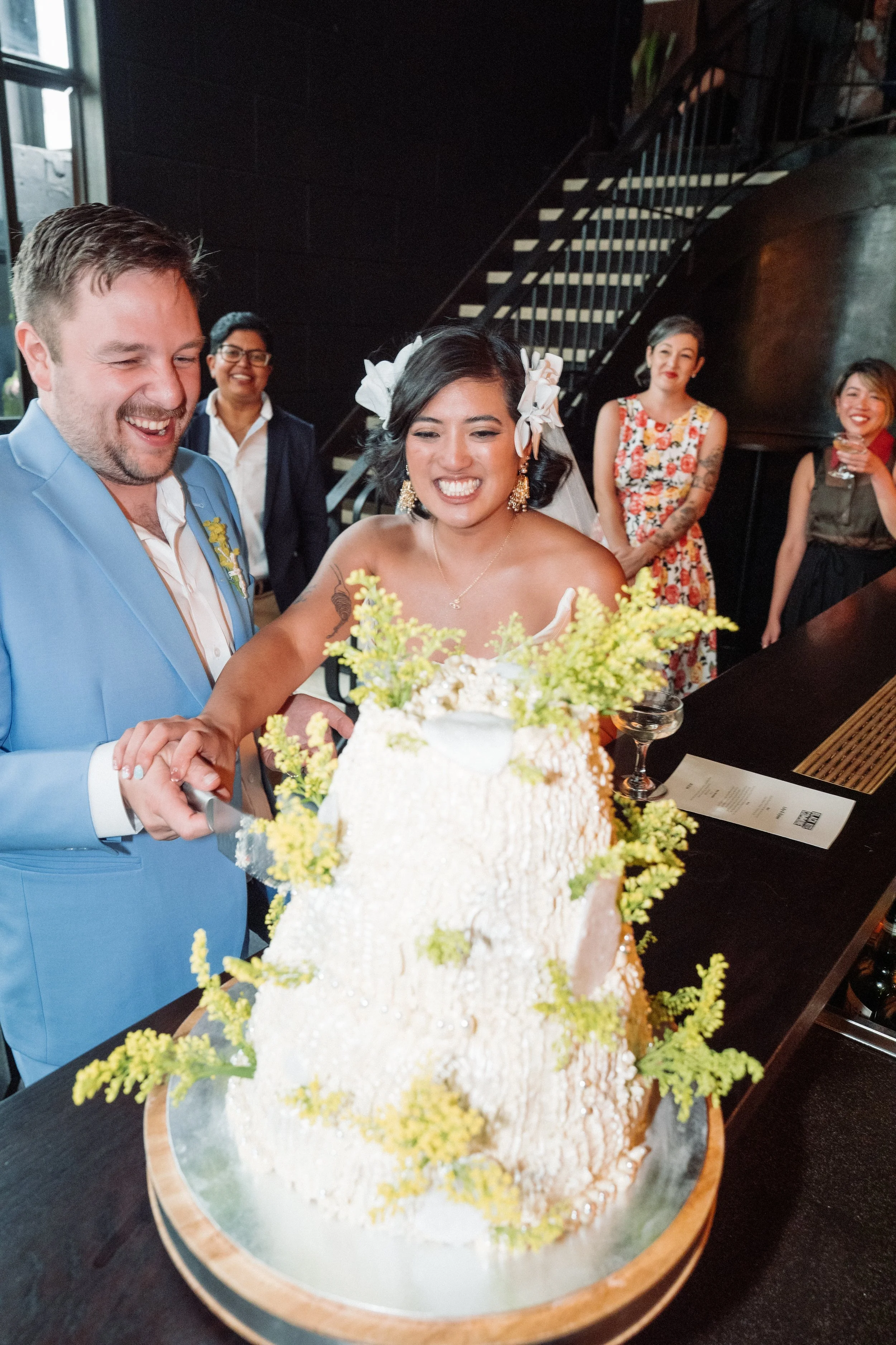 A wedding celebration where a bride and groom are cutting a wedding cake. The bride has short dark hair with flowers, and the groom is wearing a light blue suit. Behind them, four women are smiling and watching, one with glasses and tattoos, dressed 
