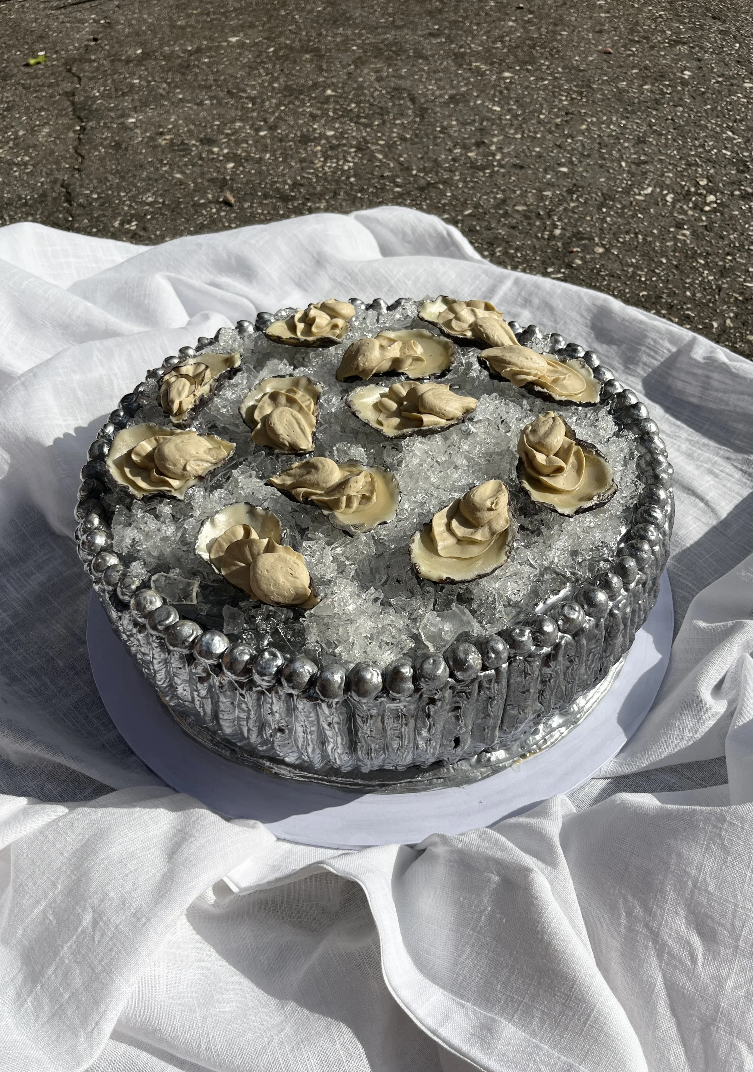 A cake made to look like a silver bowl filled with edible ice and edible chocolate oyster shells on a white tablecloth outside.