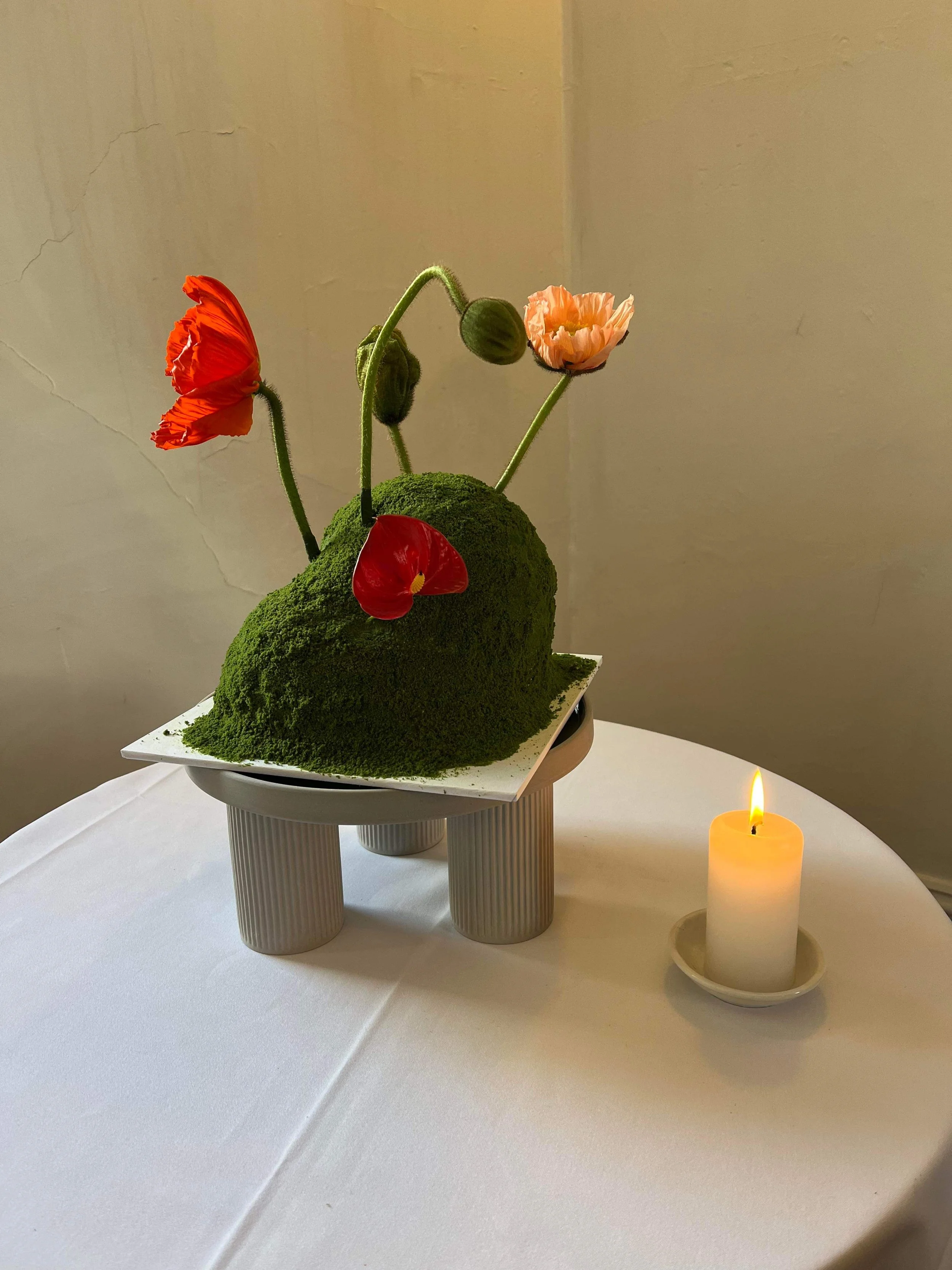 A floral arrangement featuring fresh poppies on green moss-covered mound shaped cake, placed on a white table with a lit candle nearby.