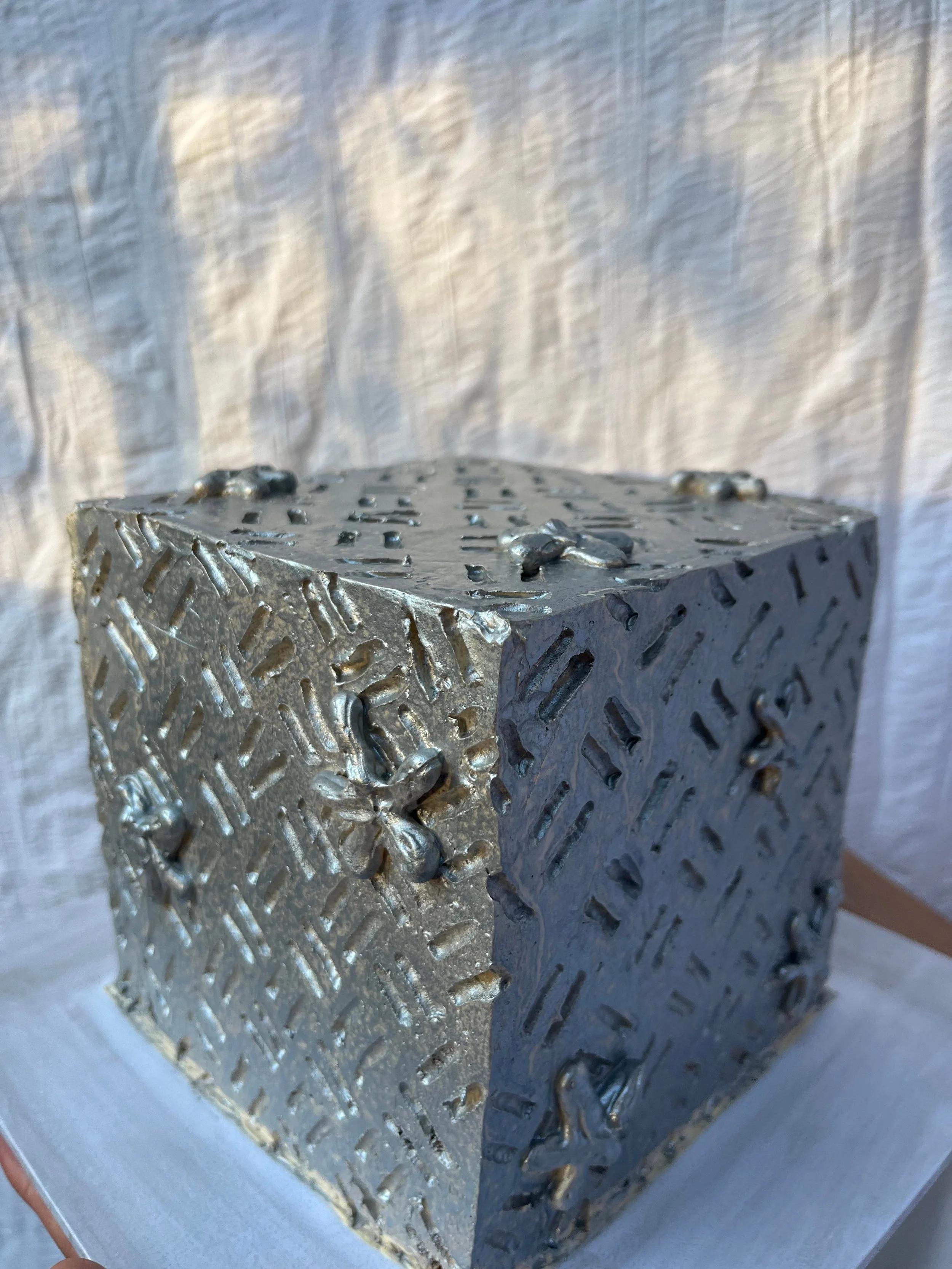 A chrome, silver, metallic, and textured cube cake with various patterns and carvings on its surface to look like metal, placed on a flat surface with a light-colored, textured fabric background.