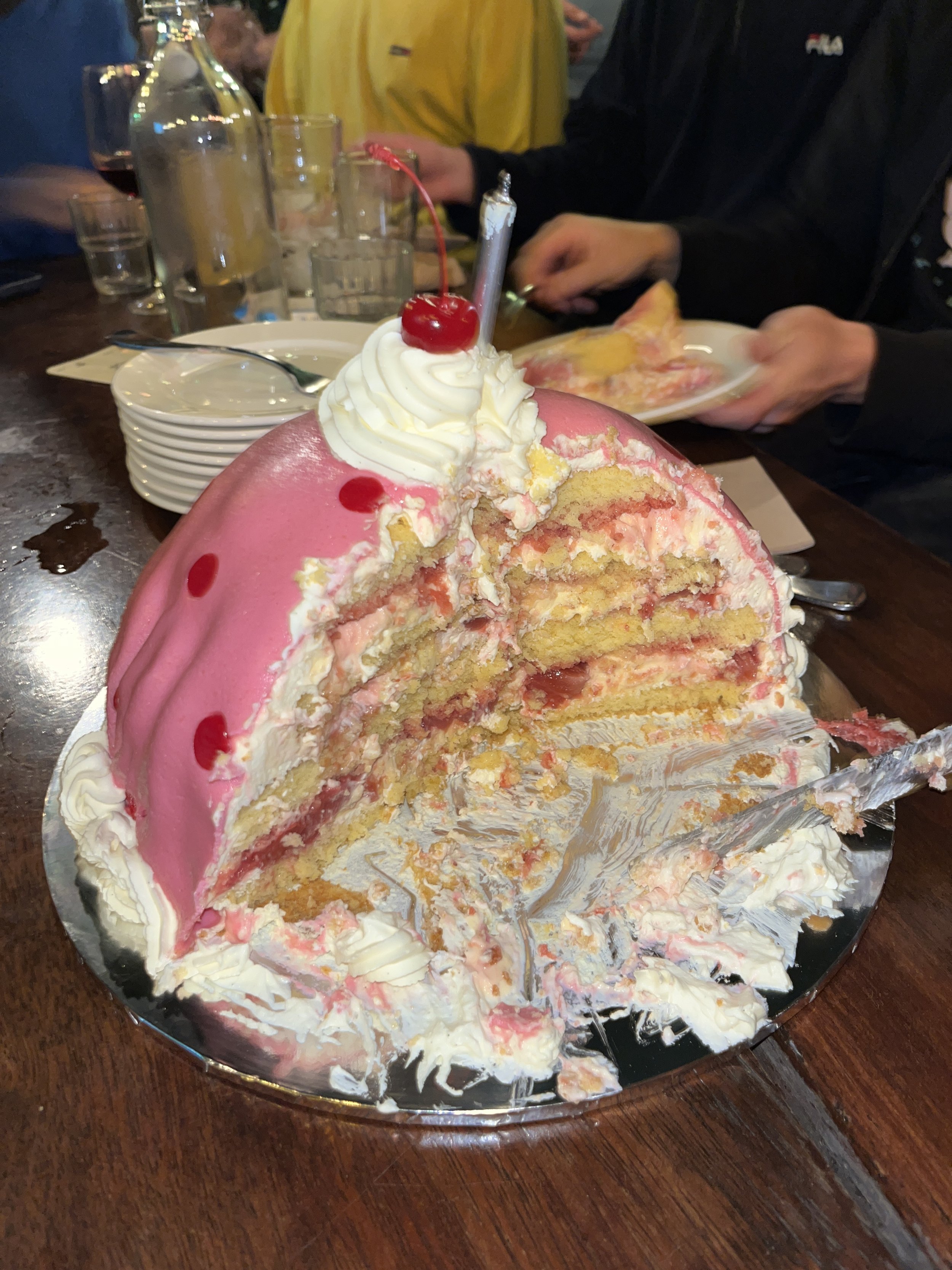 A partially eaten pink Princess Cake with red polka dots. The cake is a vanilla chiffon with strawberry jam, whipped cream, topped with a cherry, and a lit candle. The cake is on a silver platter on a wooden table.