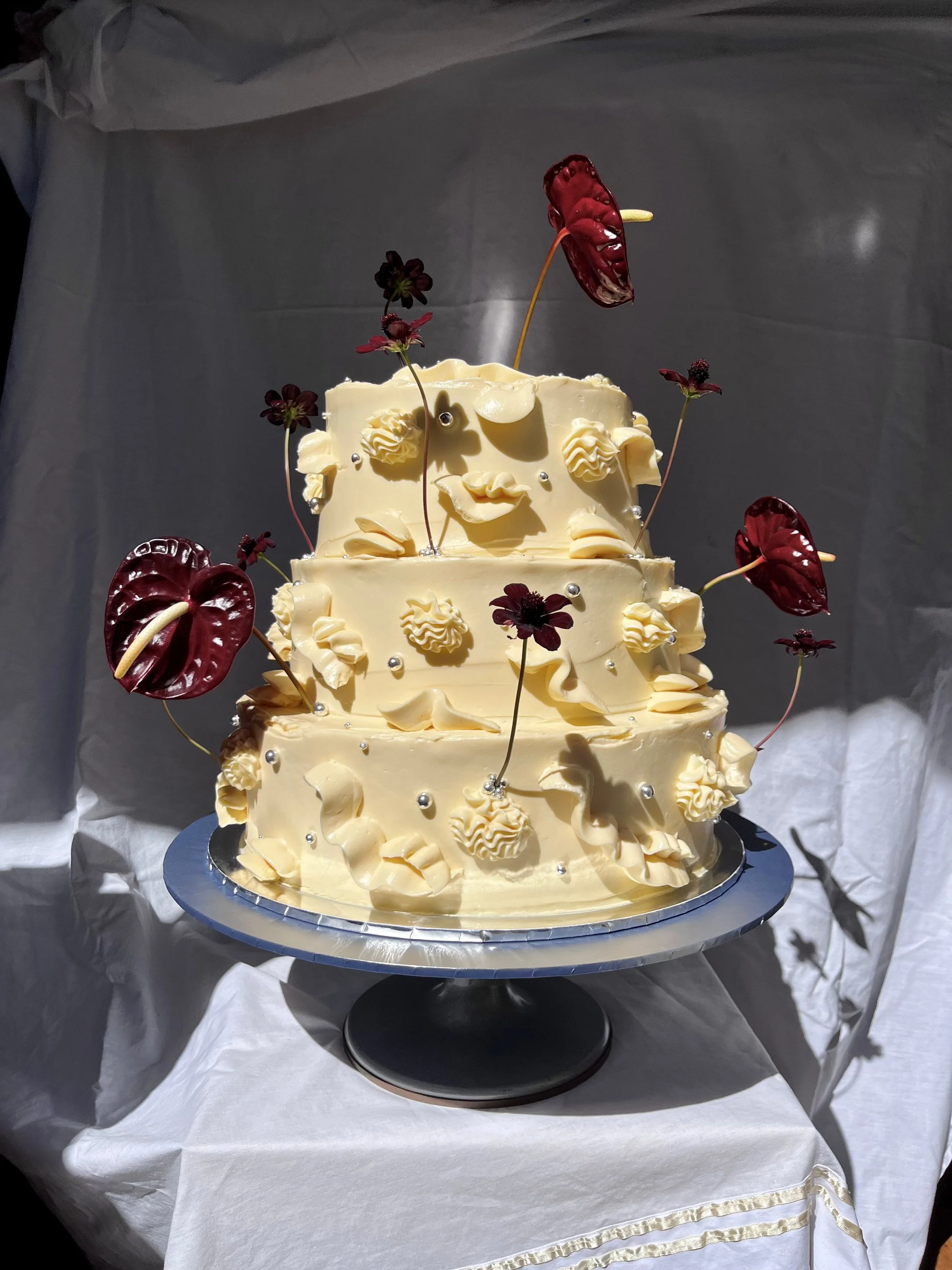 Three-tier white cake decorated with flowers and silver balls, topped with chocolate cosmos and red anthuriums, presented on a silver cake stand.