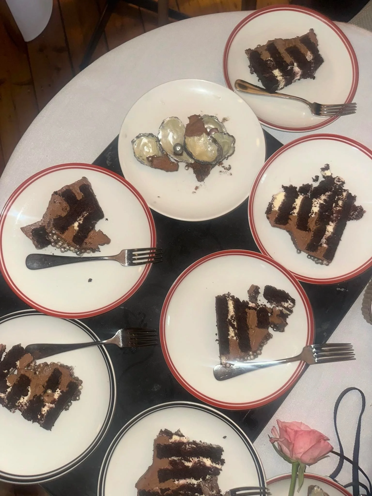 Multiple plates with slices of chocolate cake with cream filling and chocolate cream cheese frosting, some garnished with edible pearls, handpainted chocolate oysters, on a table, with a pink flower in the lower right corner.