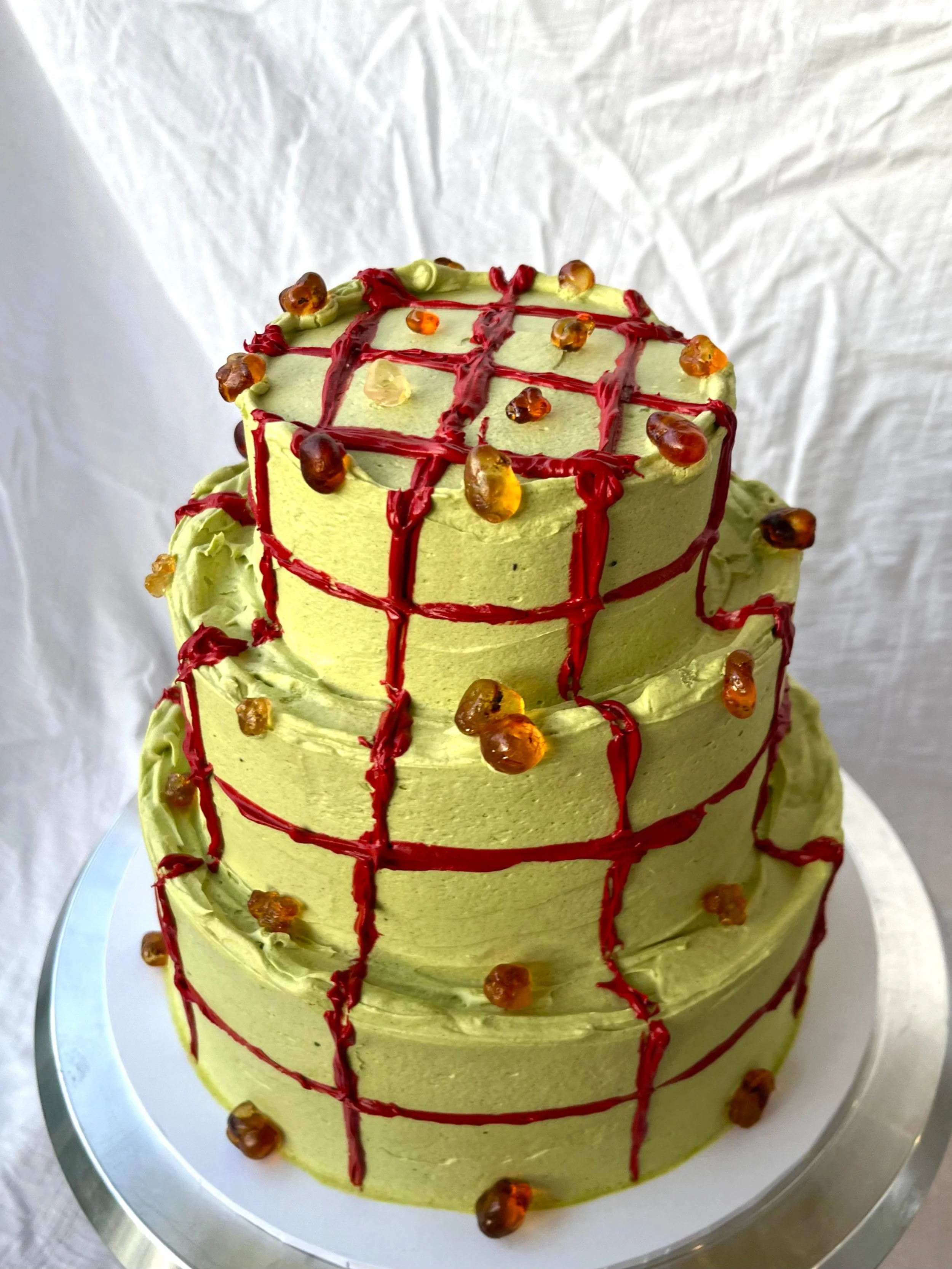 A three-tiered cake decorated with matcha frosting, topped red icing in a grid pattern and amber gemstones.