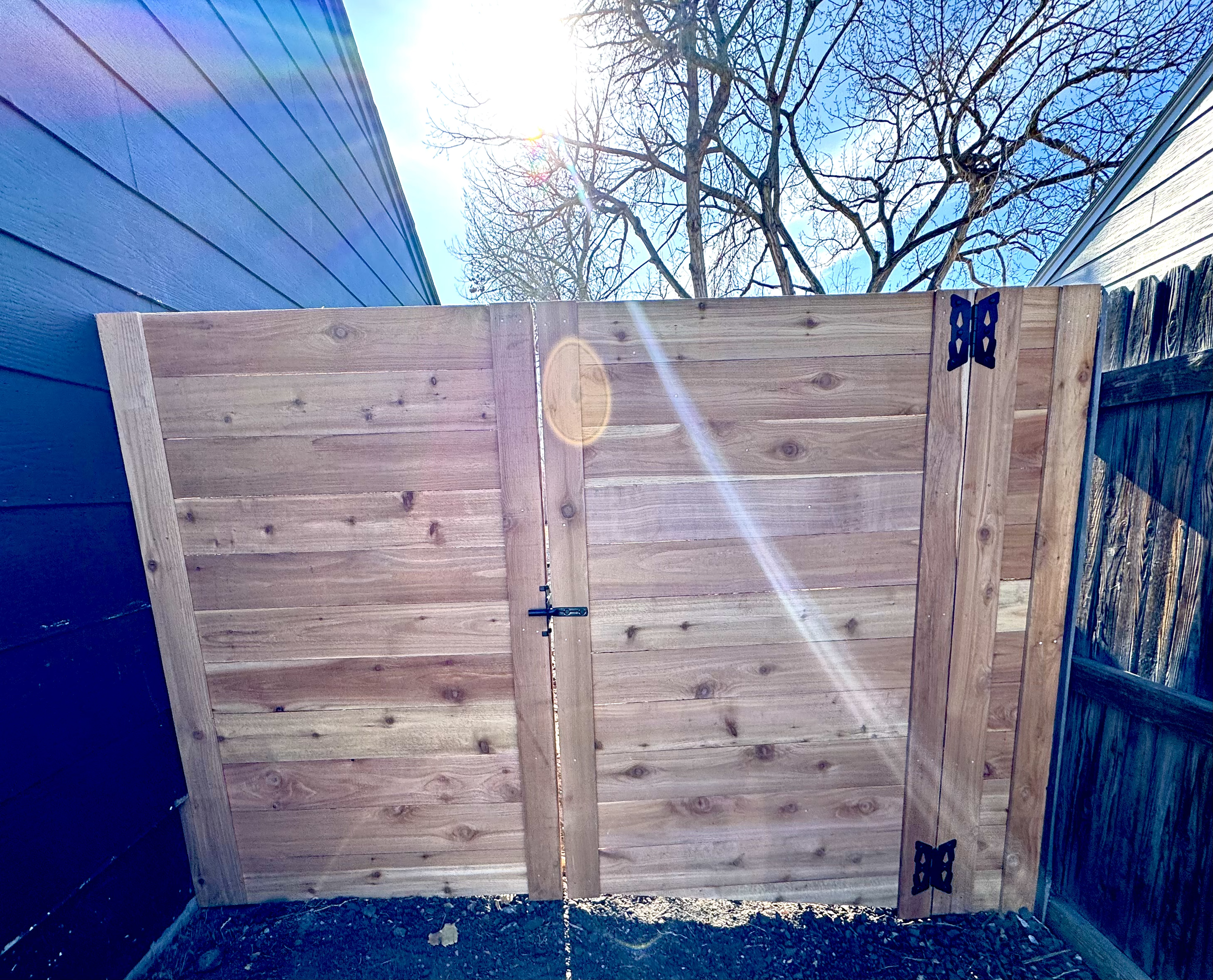 View of an outdoor wooden gate with black hinges, sunlight shining through a leafless tree in the background, clear blue sky.