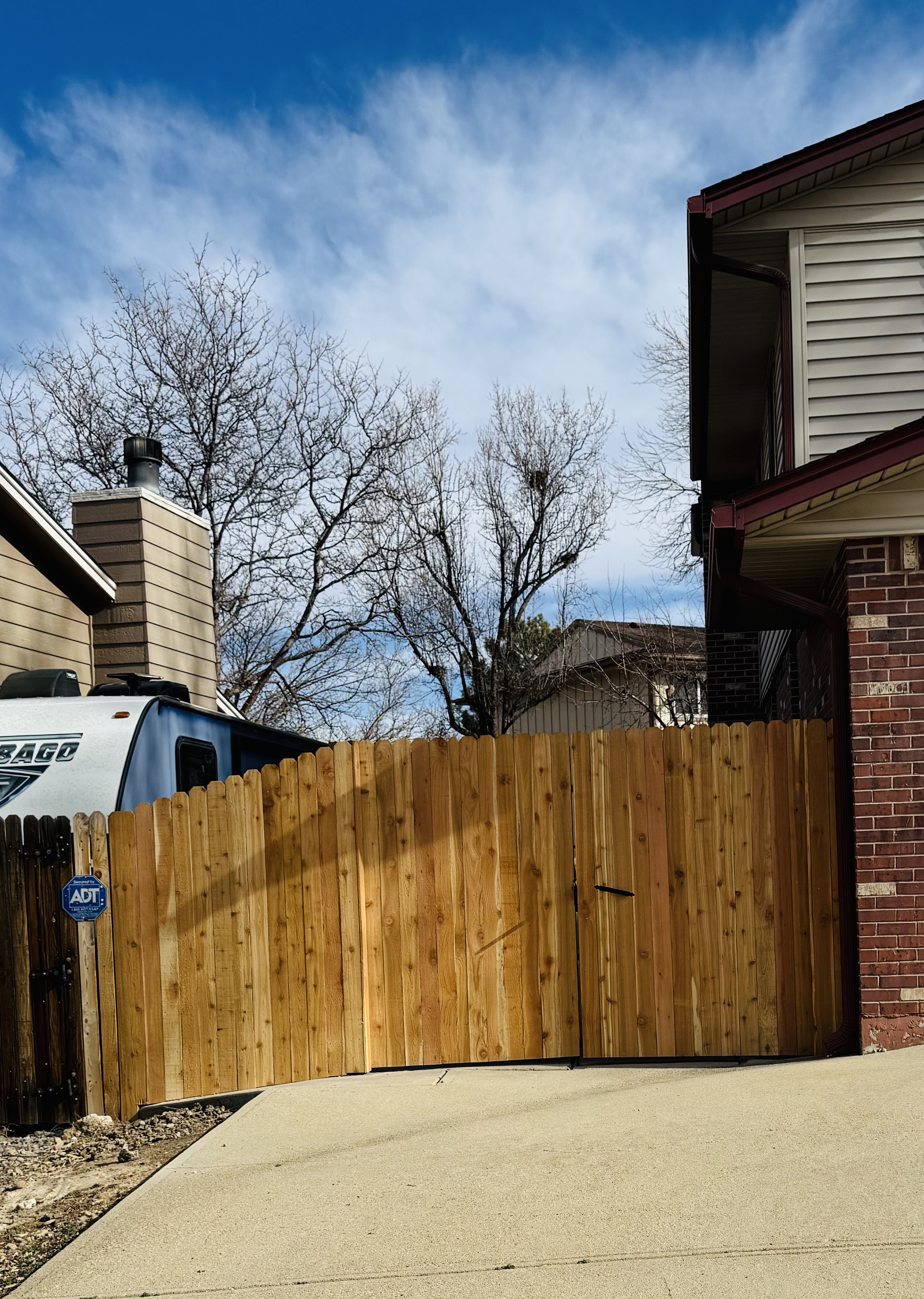 View of a wooden fence gate next to a brick house and a beige house, with leafless trees and a blue sky with clouds in the background.