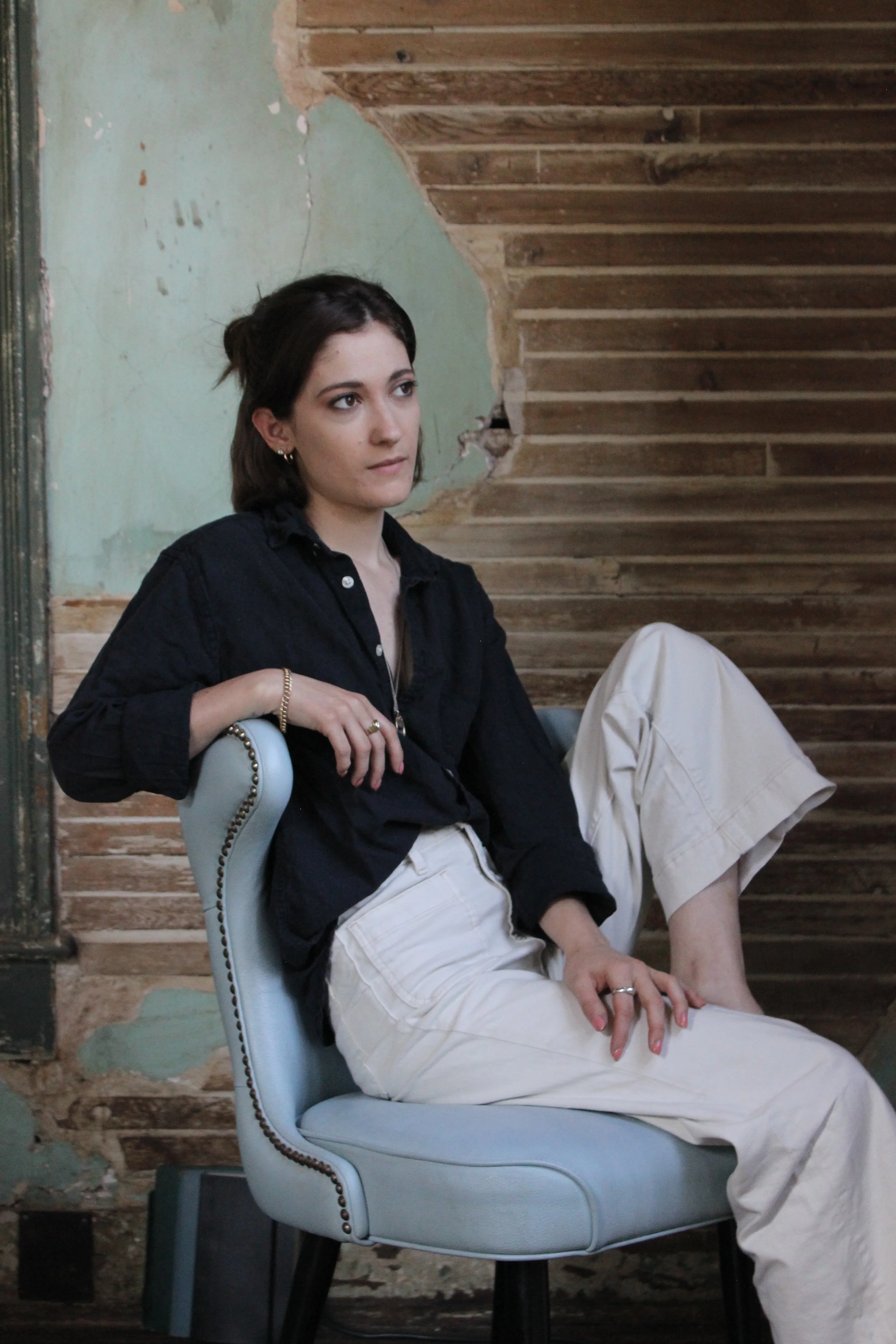 Helen Minor with dark hair, wearing a black shirt and beige pants, sitting on a light blue chair with nailhead trim, in a room with distressed green wall and exposed wooden stairs.