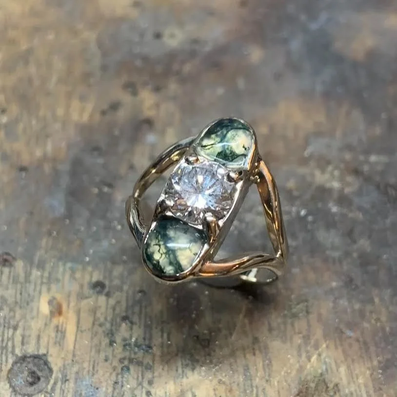A gold ring with a central square diamond and two oval-shaped greenish-blue stones on either side, set on a wooden surface.