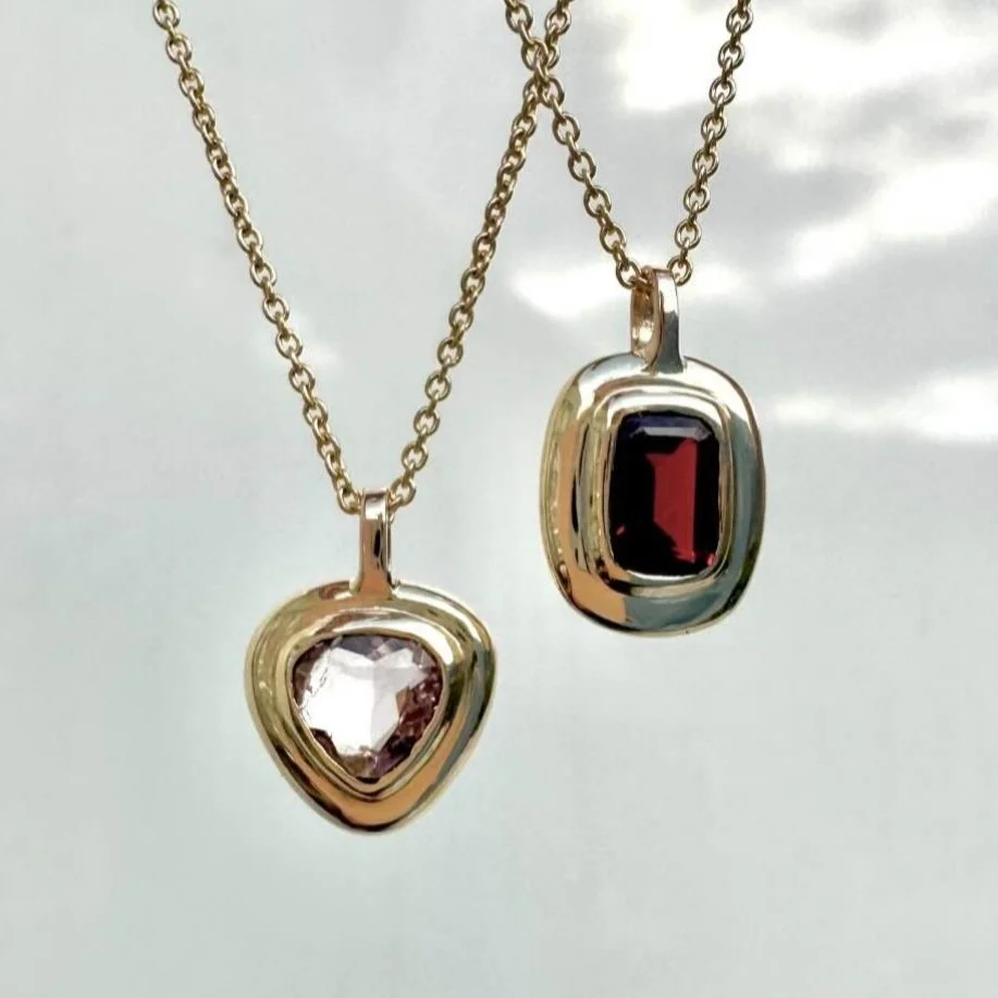 Two necklaces with gold chains, each featuring a pendant with a gemstone. One has a triangular clear gemstone, and the other has an oval red gemstone.