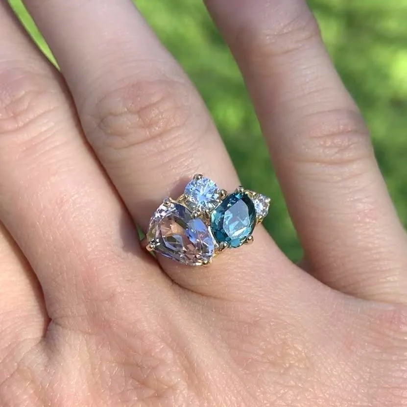 Close-up of a hand wearing a gold ring with three large gemstones in pink, blue, and clear, set on a green background.