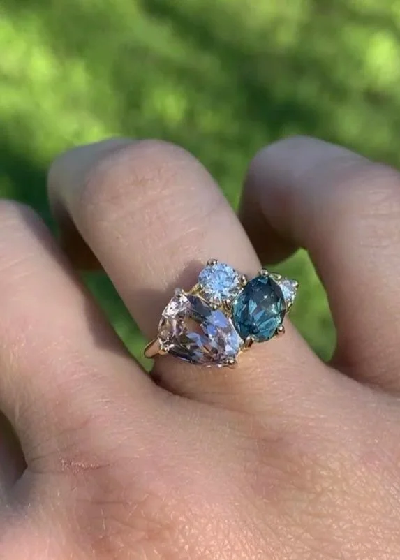 Close-up of a hand wearing a large ring with three prominent stones, one blue and two clear, set against a blurred green background.