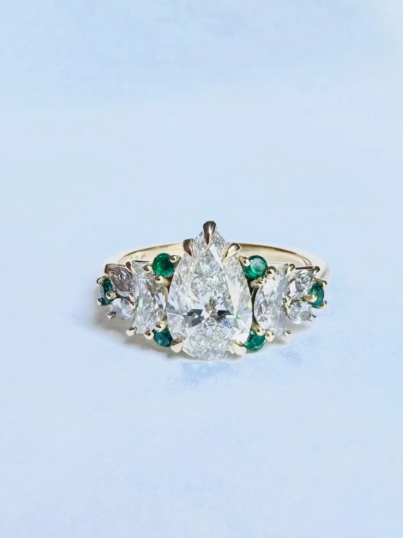A silver ring with a large pear-shaped diamond at the center, surrounded by smaller diamonds and green gemstones.