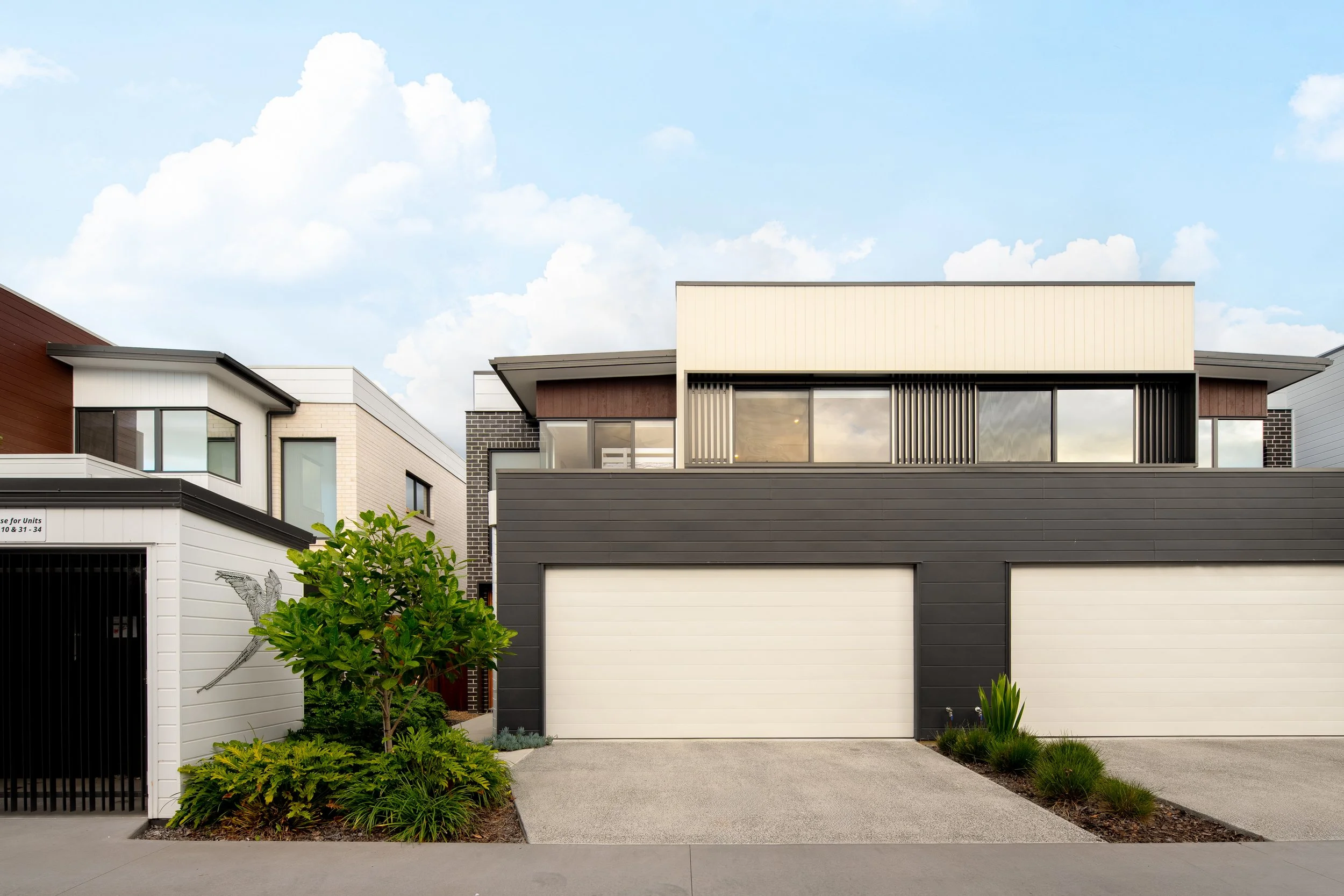 Modern residential building with a white garage door, black and white siding, and greenery in front, under a partly cloudy sky.