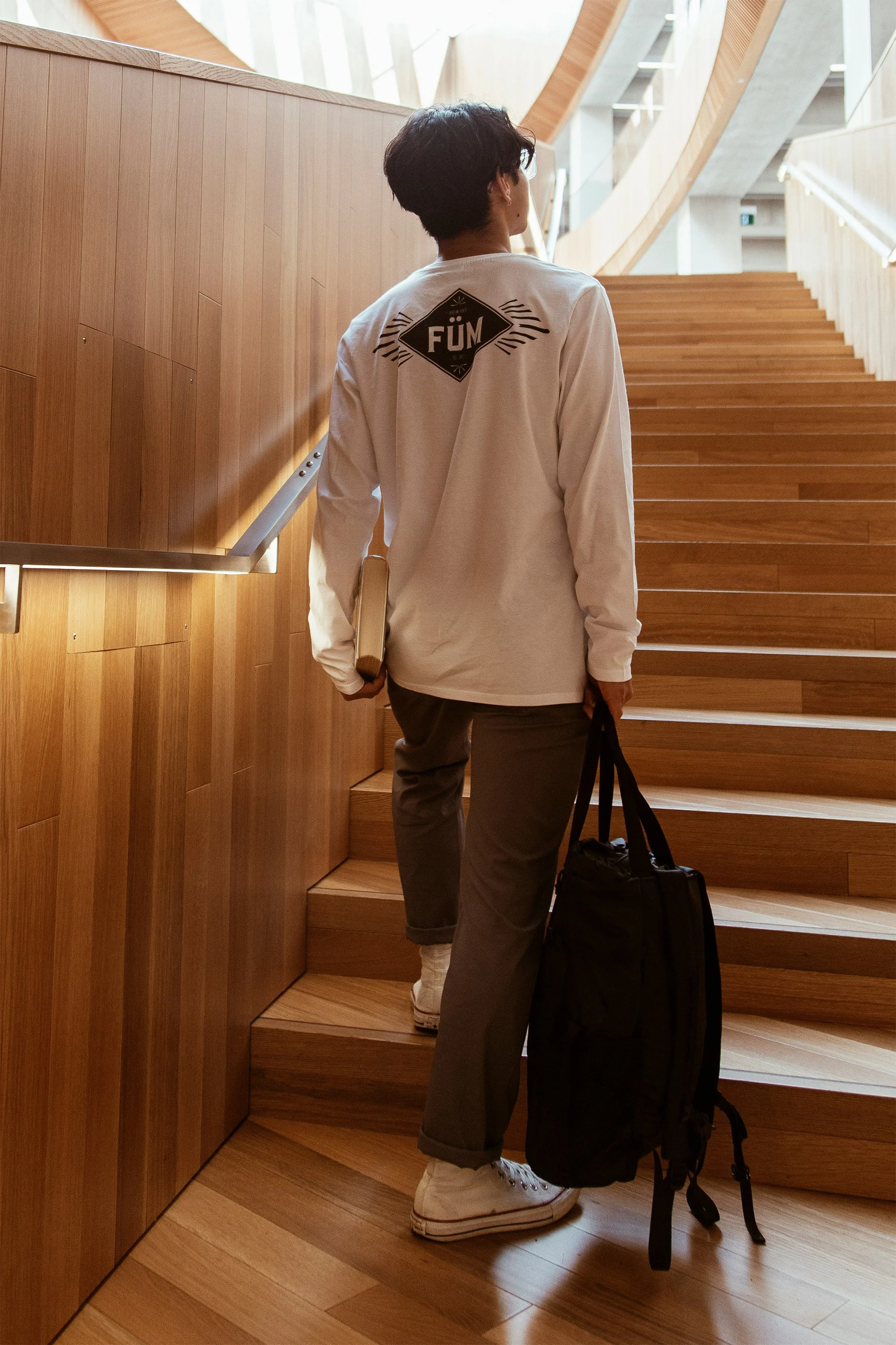 A person with short dark hair wearing glasses, a white long-sleeve shirt with a design on the back, gray pants, and white sneakers, standing on wooden stairs inside a modern building with wooden and concrete architectural elements, carrying a black bag and holding a white object, possibly a folder or notebook.