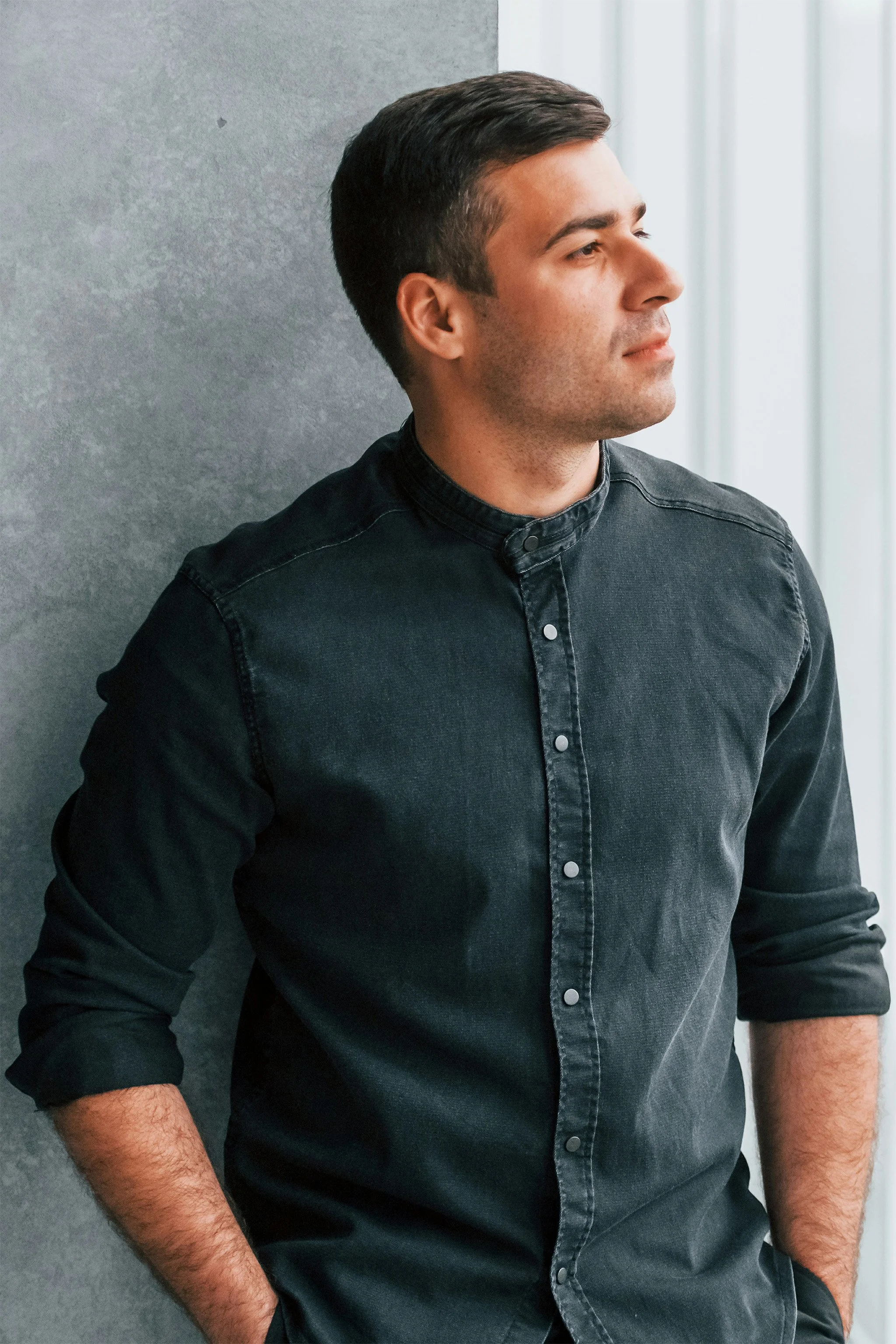 Side profile of a man with dark hair wearing a black button-up shirt, standing against a gray wall.