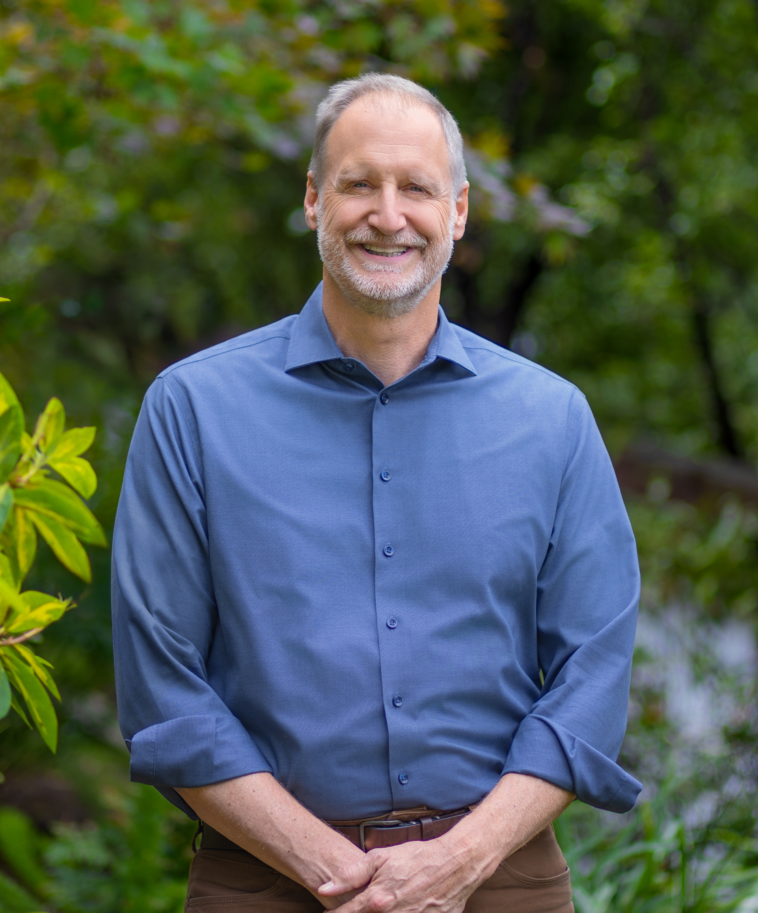 A smiling middle-aged man with gray hair and a beard wearing a blue dress shirt, standing outdoors in front of blurred green foliage.