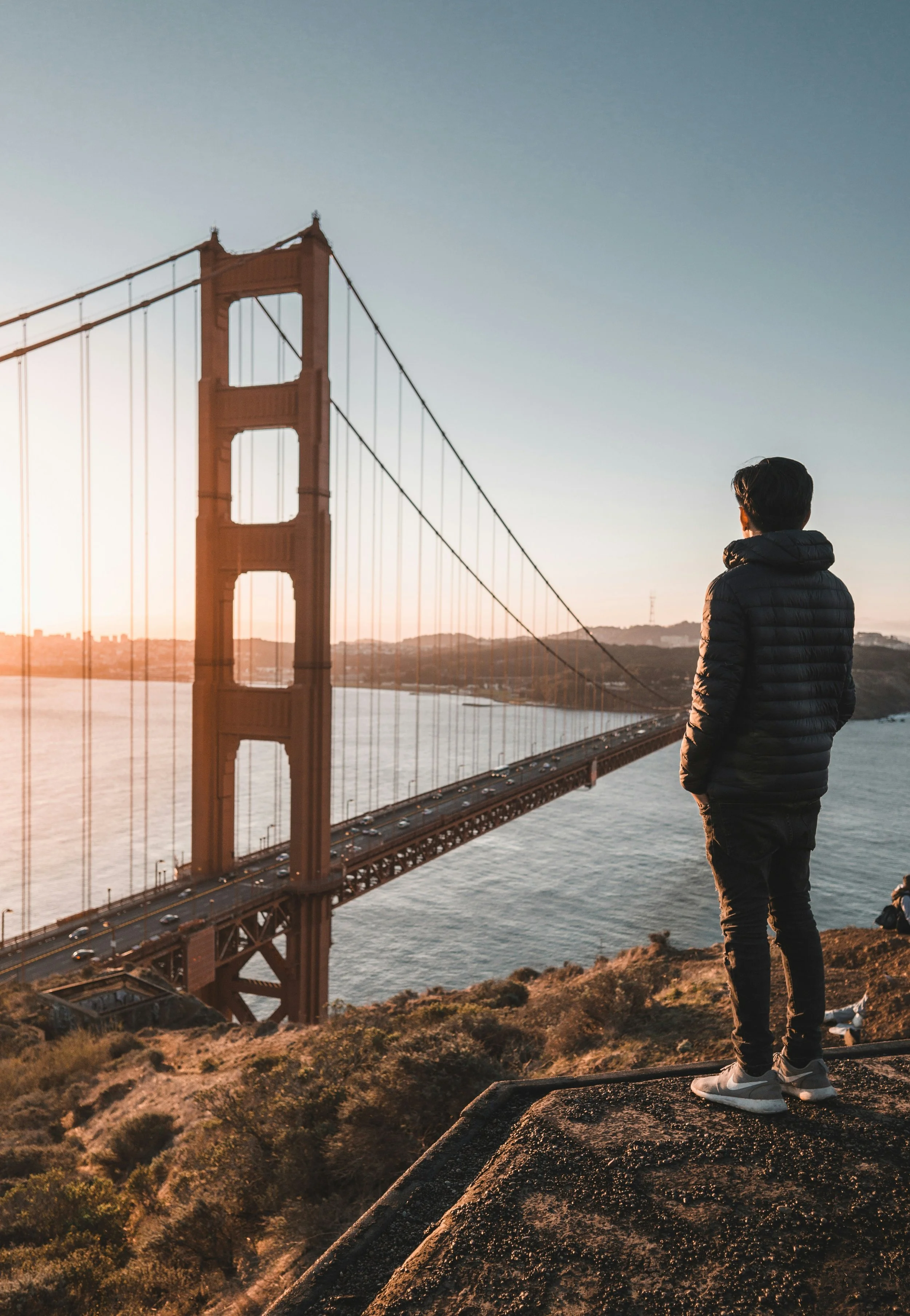 A man in a black jacket and gray sneakers standing on a ledge overlooking the Golden Gate Bridge at sunset.