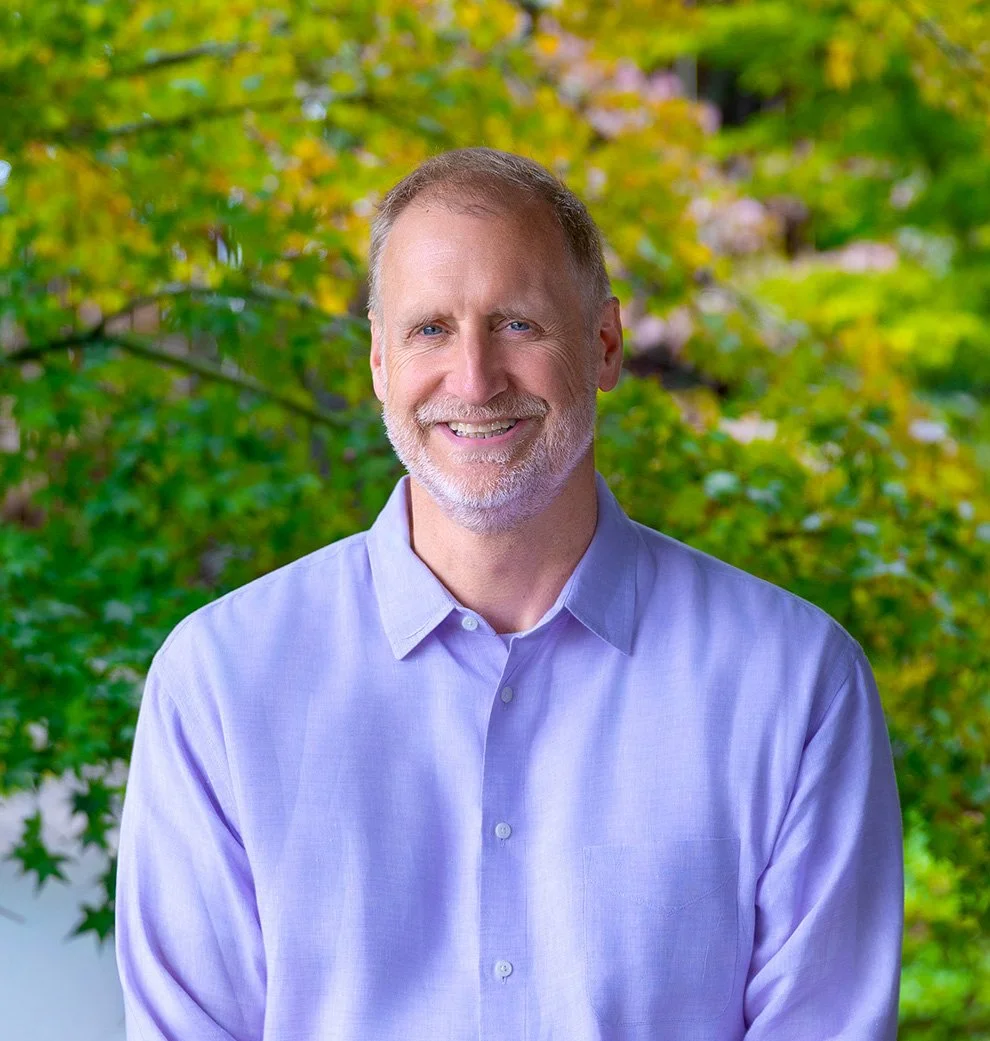 A smiling middle-aged man with a beard and mustache wearing a light purple dress shirt, outdoors with green and yellow autumn leaves in the background.