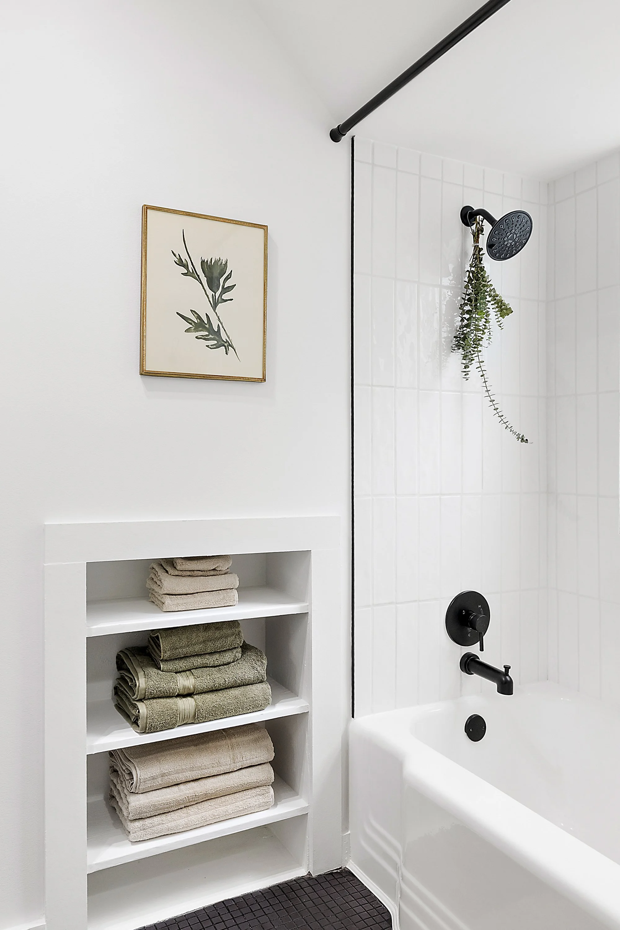 A renovated bathroom with a built in cabinet, tub, black shower head, green and tan towels with a picture of a leaf on the wall. 
