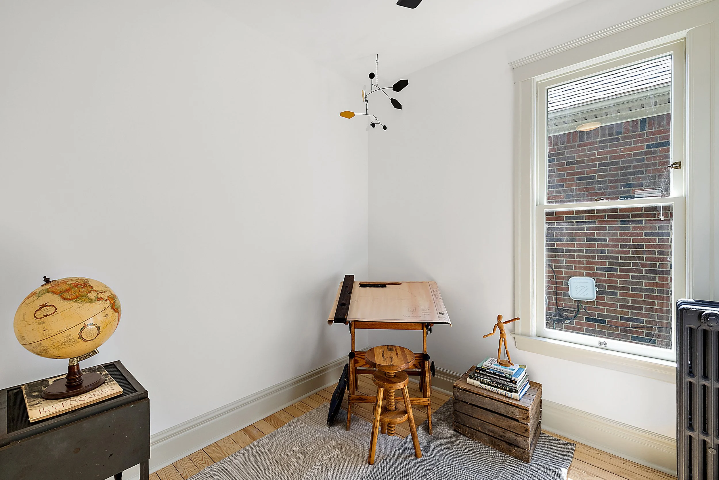 Renovated room with white walls, a window, carpets on the floor, a mobile hanging from the ceiling, and a drawing table. 