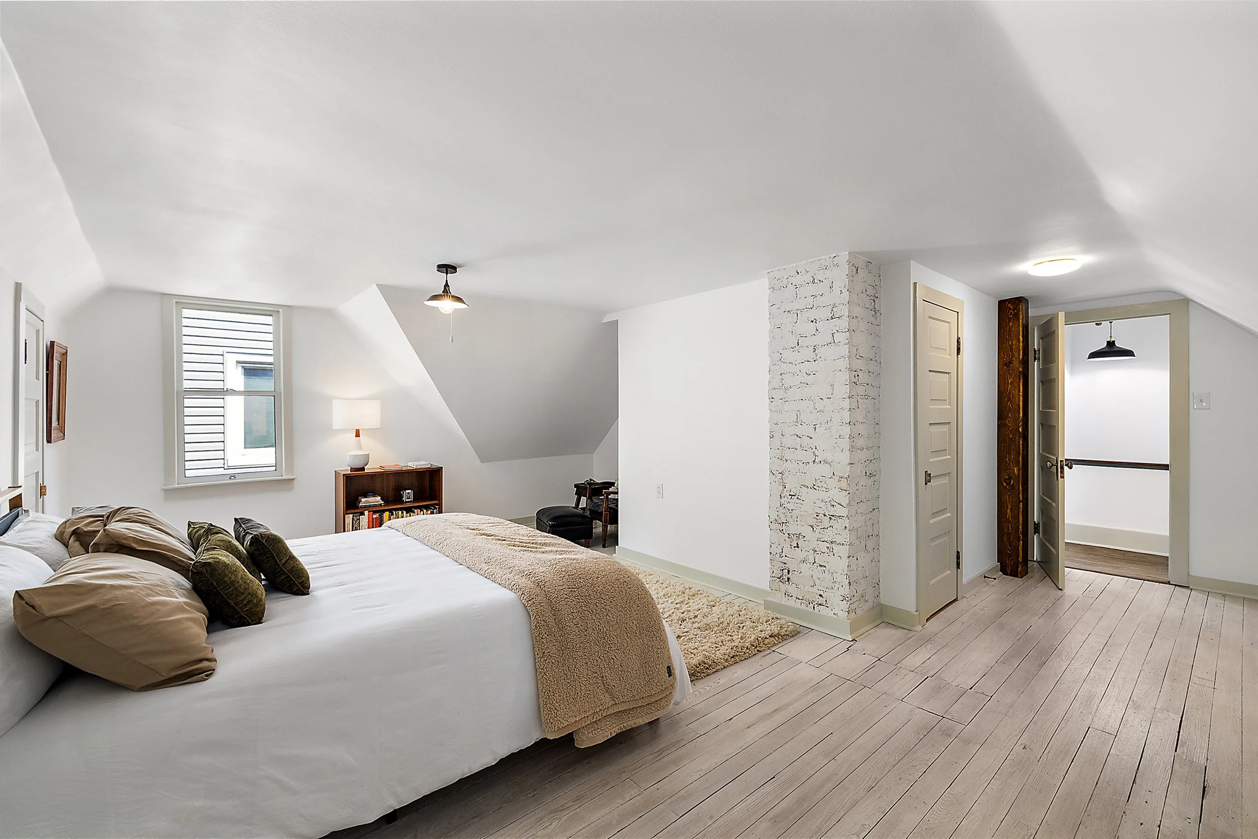 A renovated bedroom with renovated floors and walls. In the room there is a window, door, and bookshelf. There is a pendant light and a lamp along with a chimney and closet. 