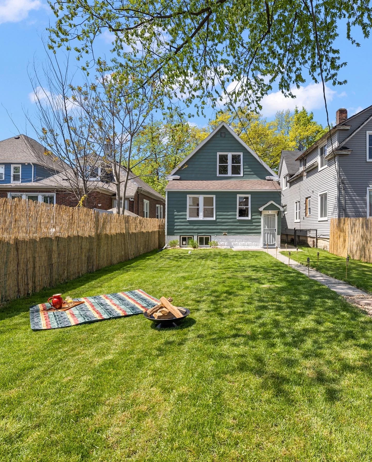 A 1915 Edwardian house that is green with a beautiful backyard with grass, a fire pit, and a picnic blanket. 