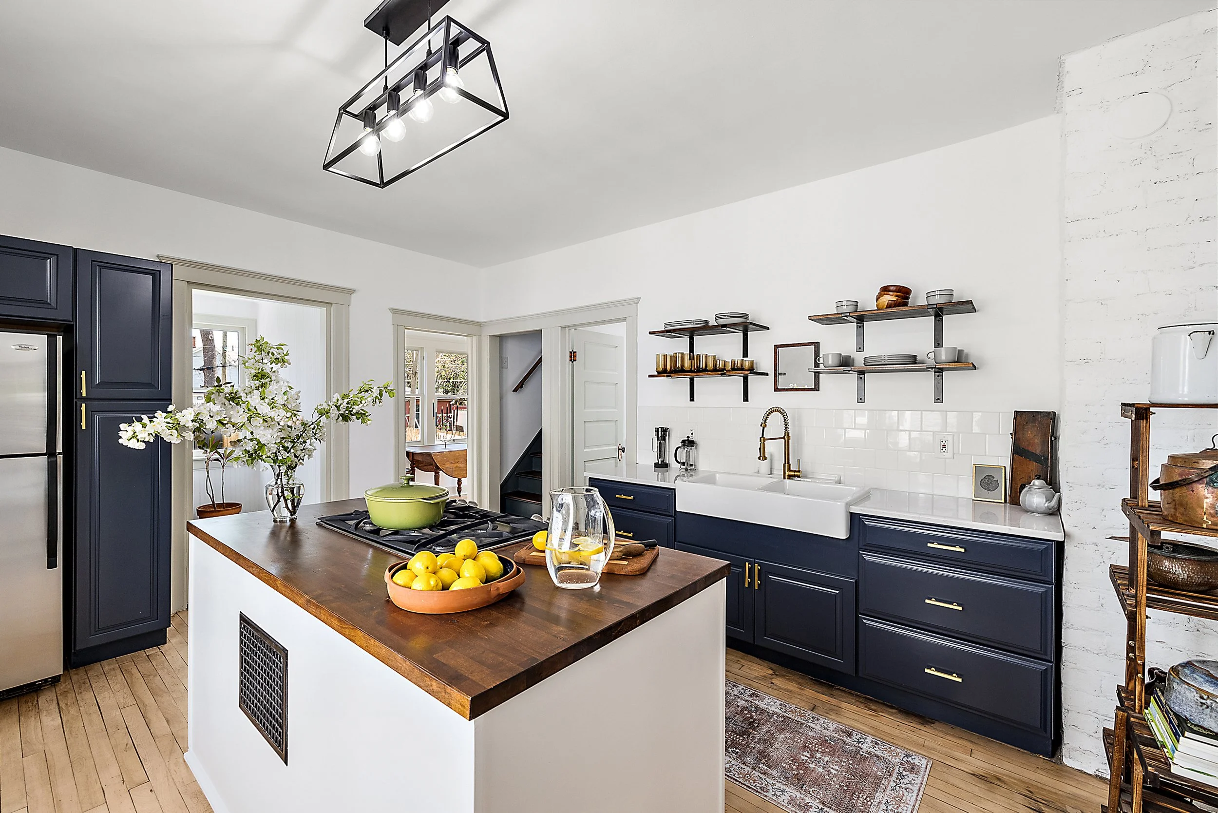 Renovation kitchen with home decor on butcher block island next to blue cabinets, white farmhouse style sink and custom wood shelves.