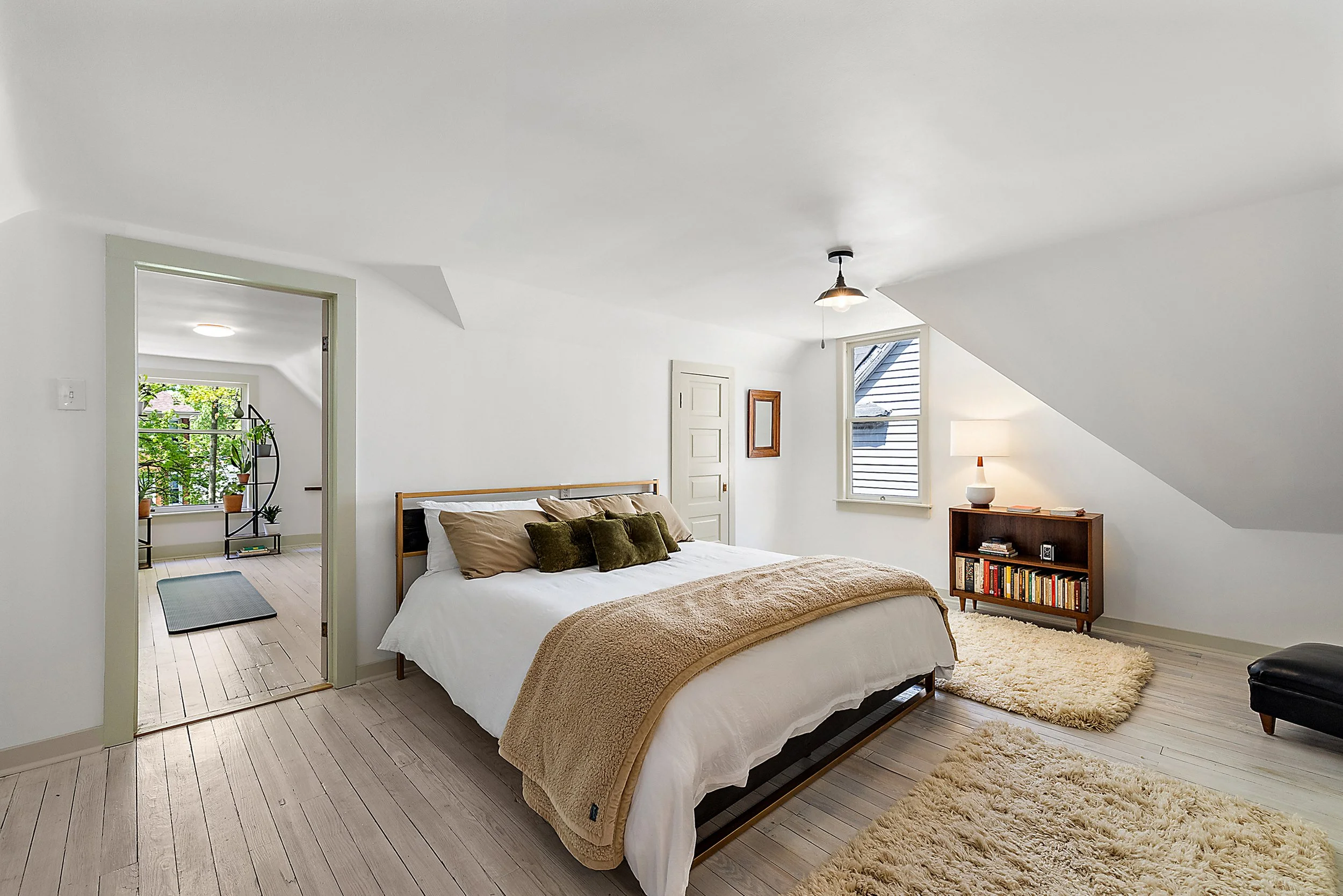 A renovated bedroom with renovated floors and walls. In the room there is a window, door, and bookshelf. There is a pendant light and a lamp. 