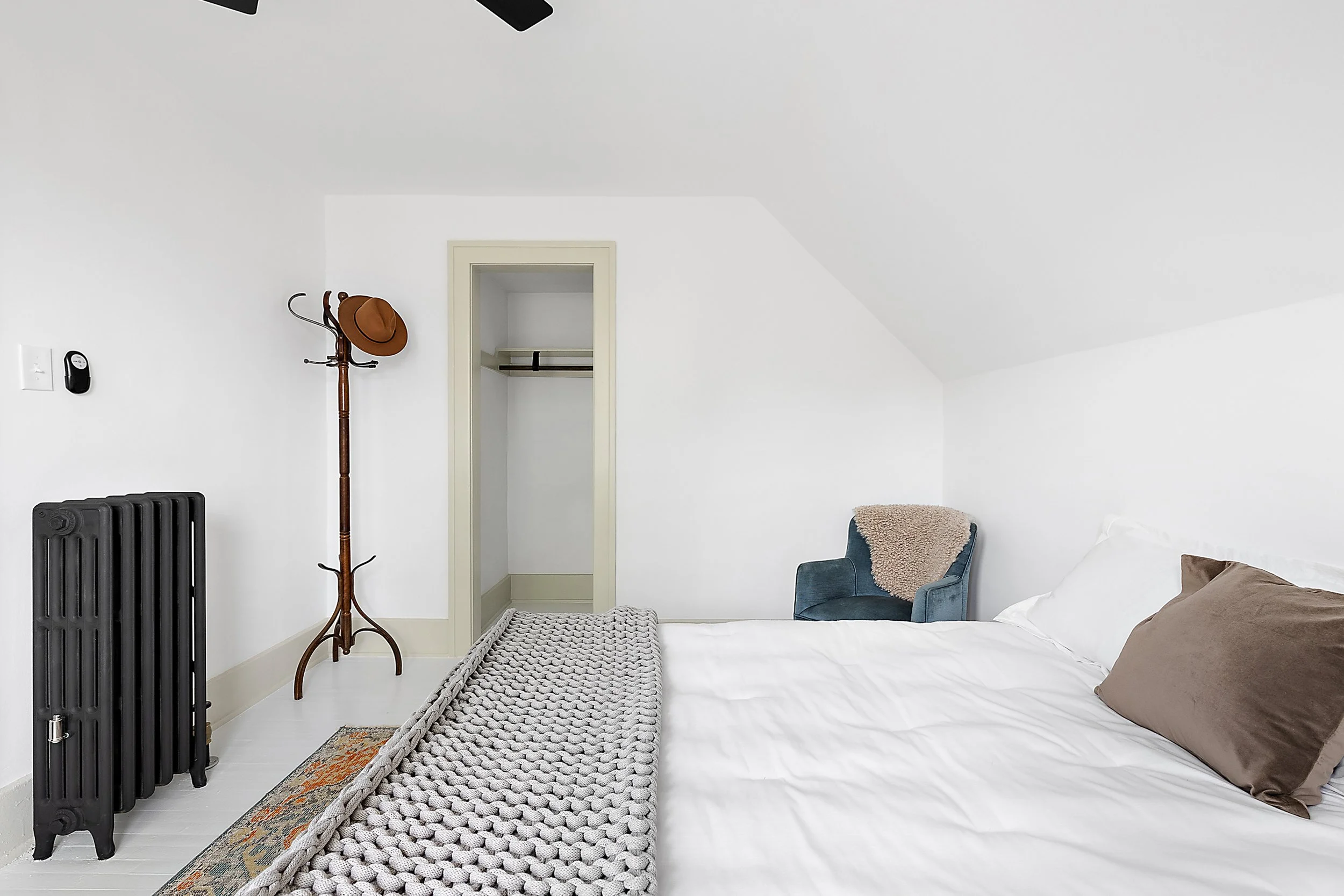 A renovated bedroom with a black radiator, white plaster walls, a bed, a closet with green trim and a coat rack and hat. 