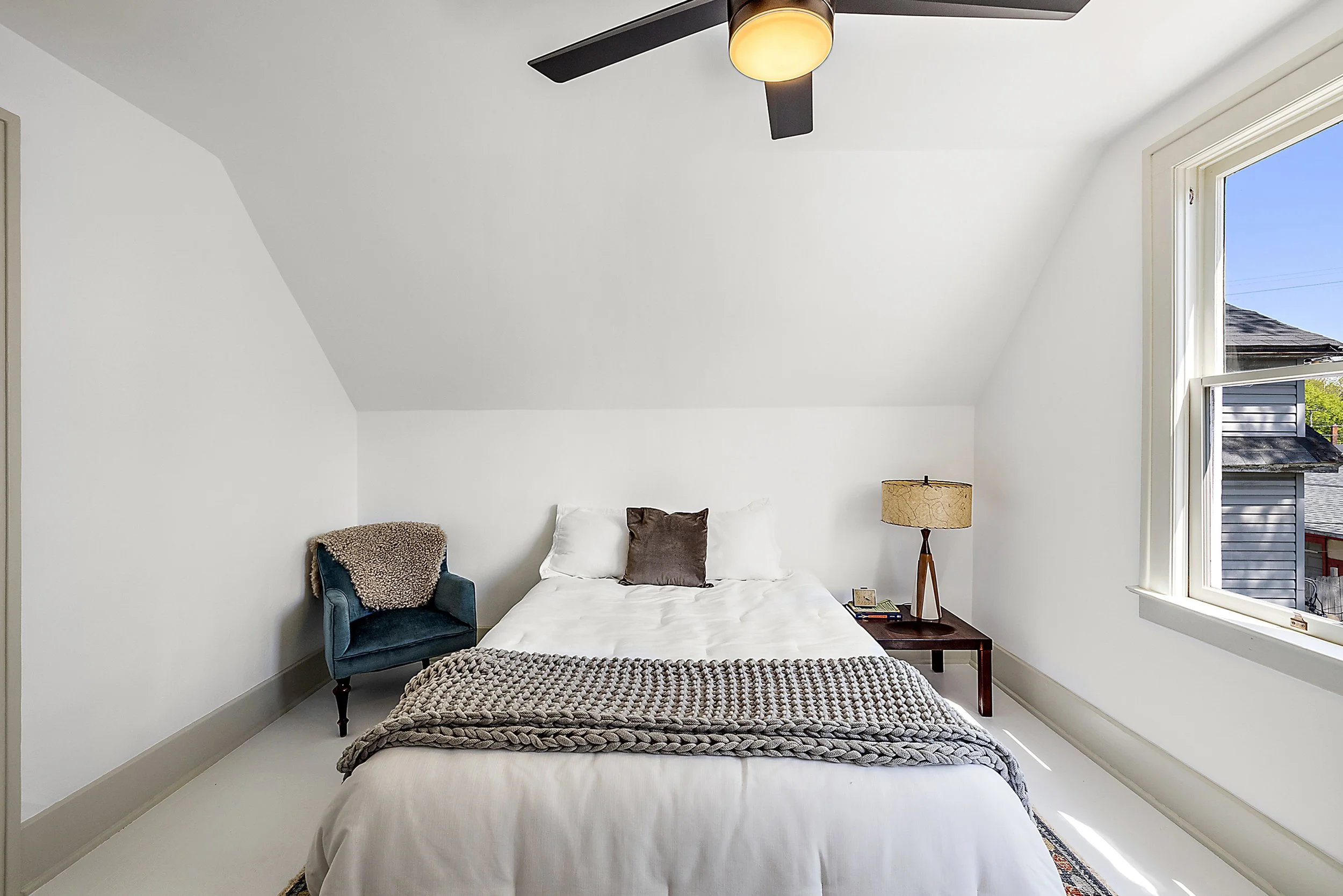 A renovated bedroom with a bed, chair, white walls, and side table with a lamp. 